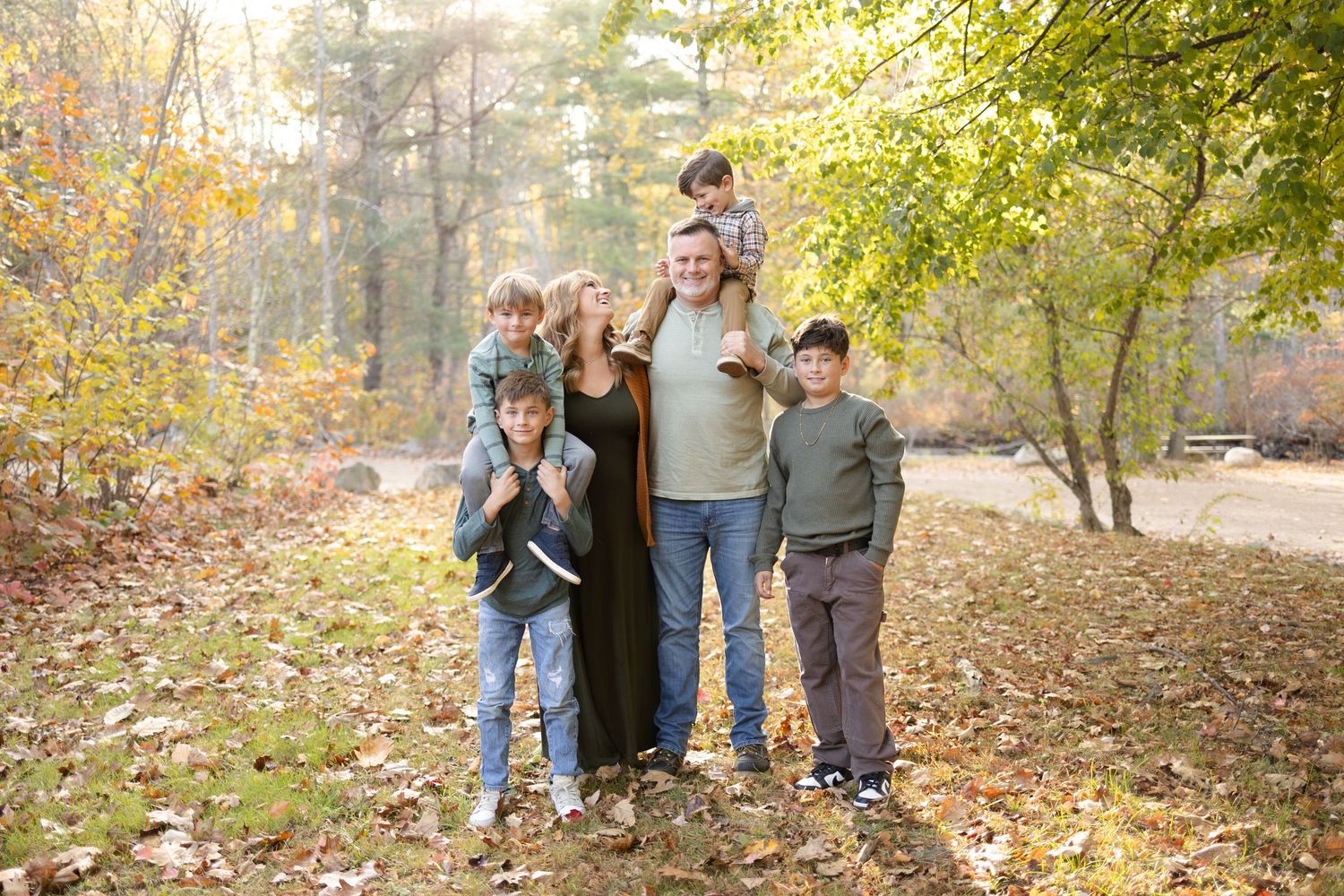 Family of six poses among autumn leaves in a wooded park, with children on shoulders and smiling faces.