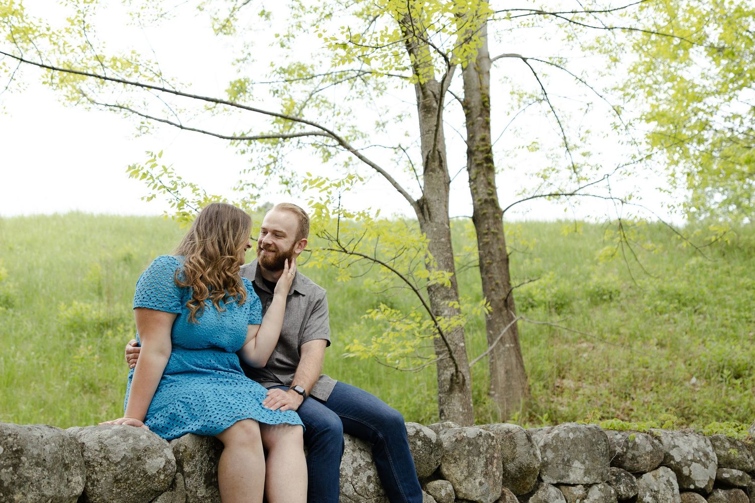 Couple sitting on a stone wall in a lush green field, sharing a romantic moment near blooming trees.