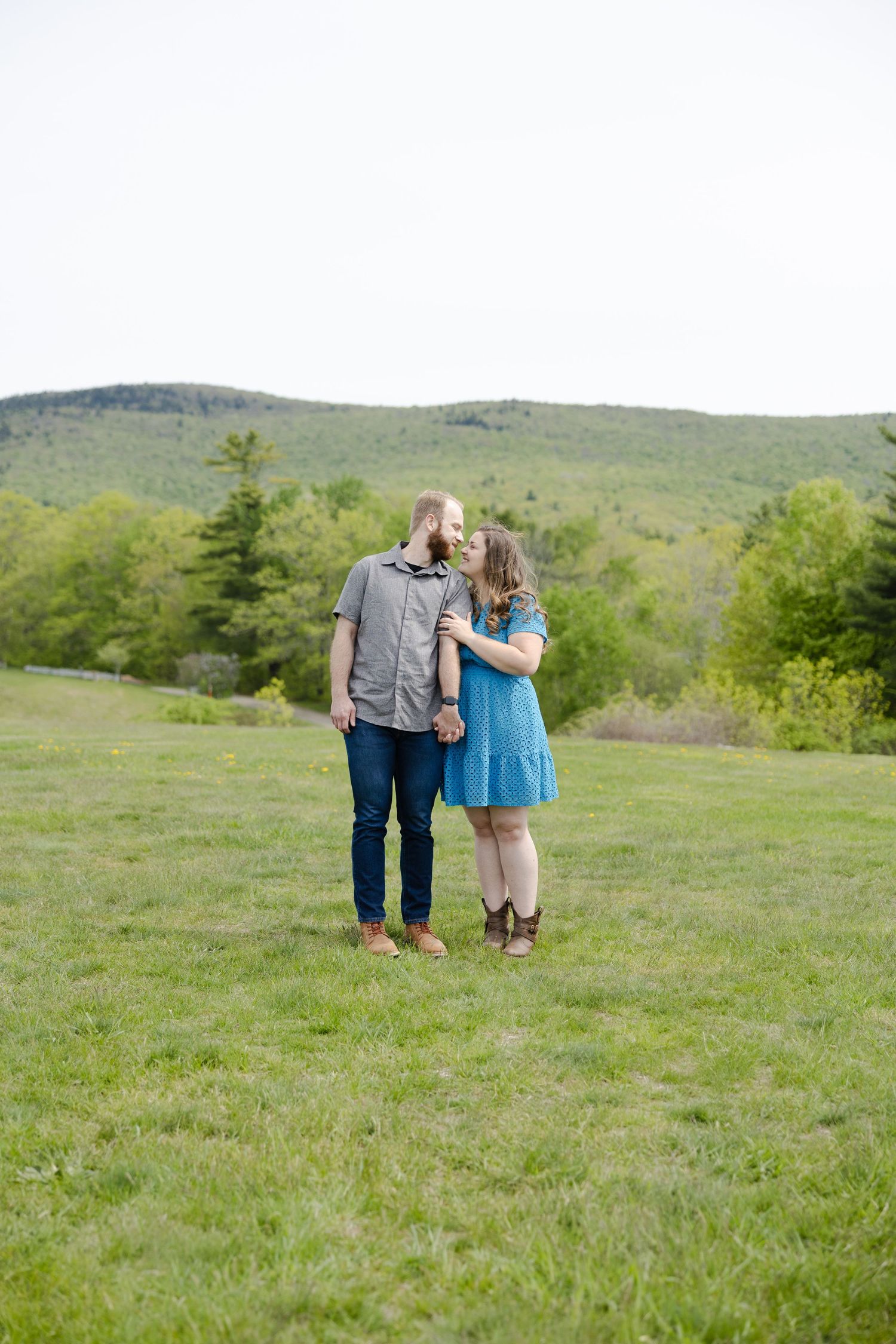 Couple sharing a kiss in a green meadow with rolling forested hills in the background on a cloudy day.