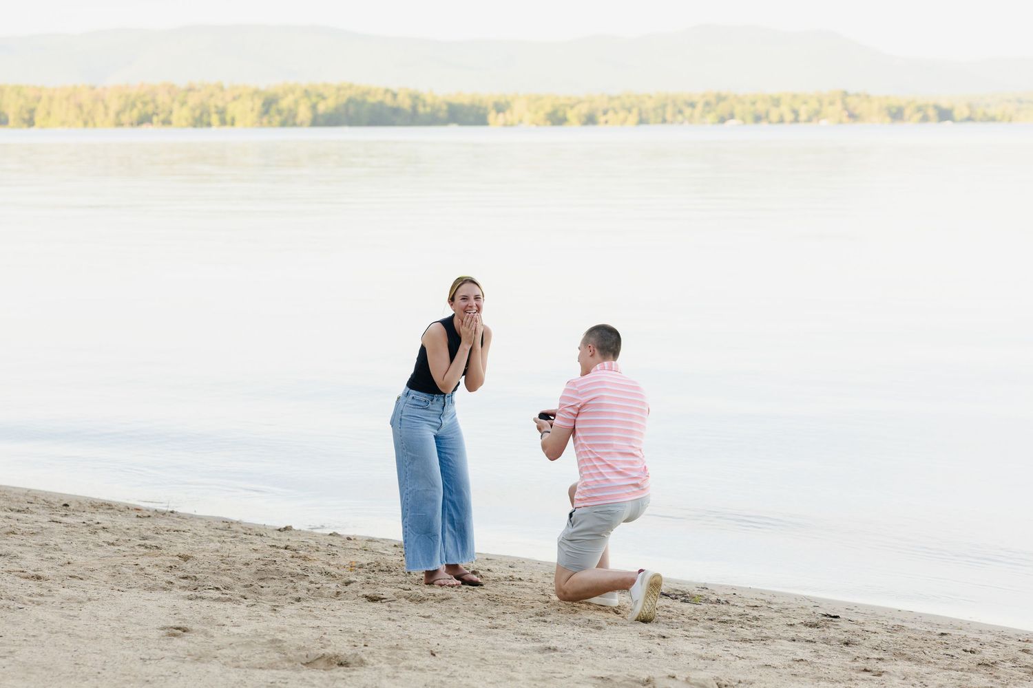 Man proposing on a sandy beach at sunset while woman reacts with surprise, with a serene lake and mountains behind.
