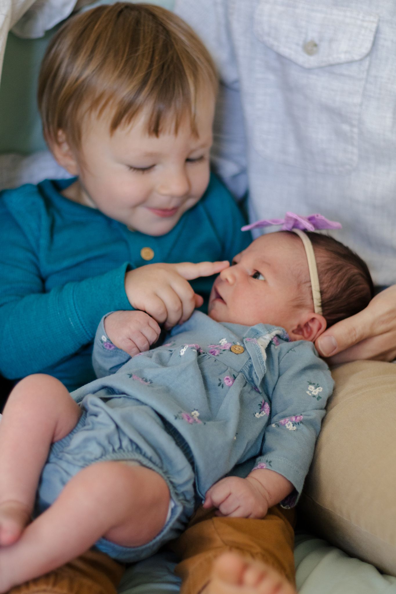 A child in a teal sweater gently touches a baby wearing a floral outfit and headband while resting on cushions.