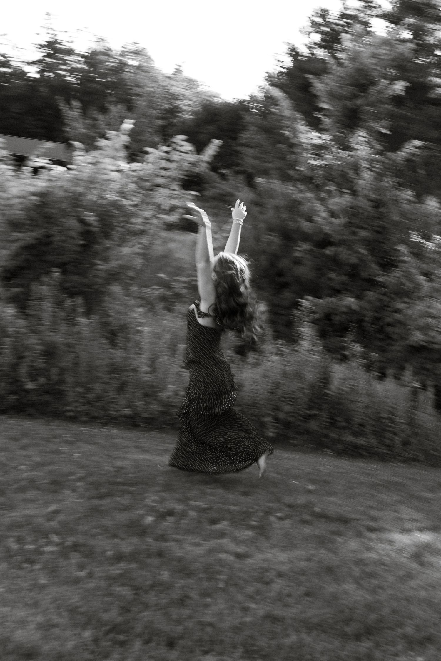 Black and white photo of a woman in a dress jumping joyfully with arms raised in a lush green garden.