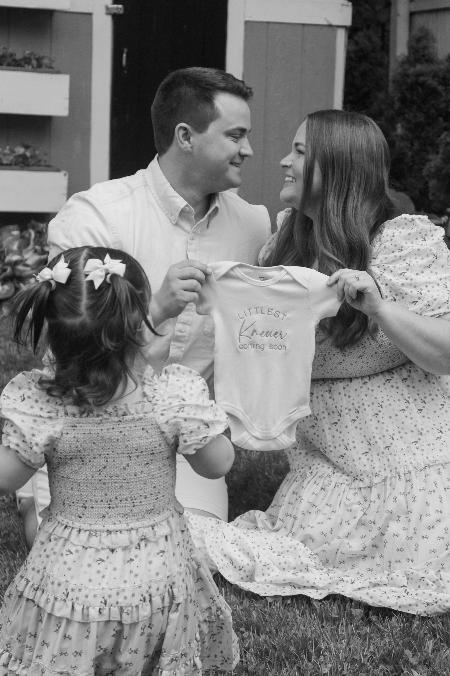 Black and white photo of a couple holding a baby onesie while their young daughter looks on, announcing a pregnancy.
