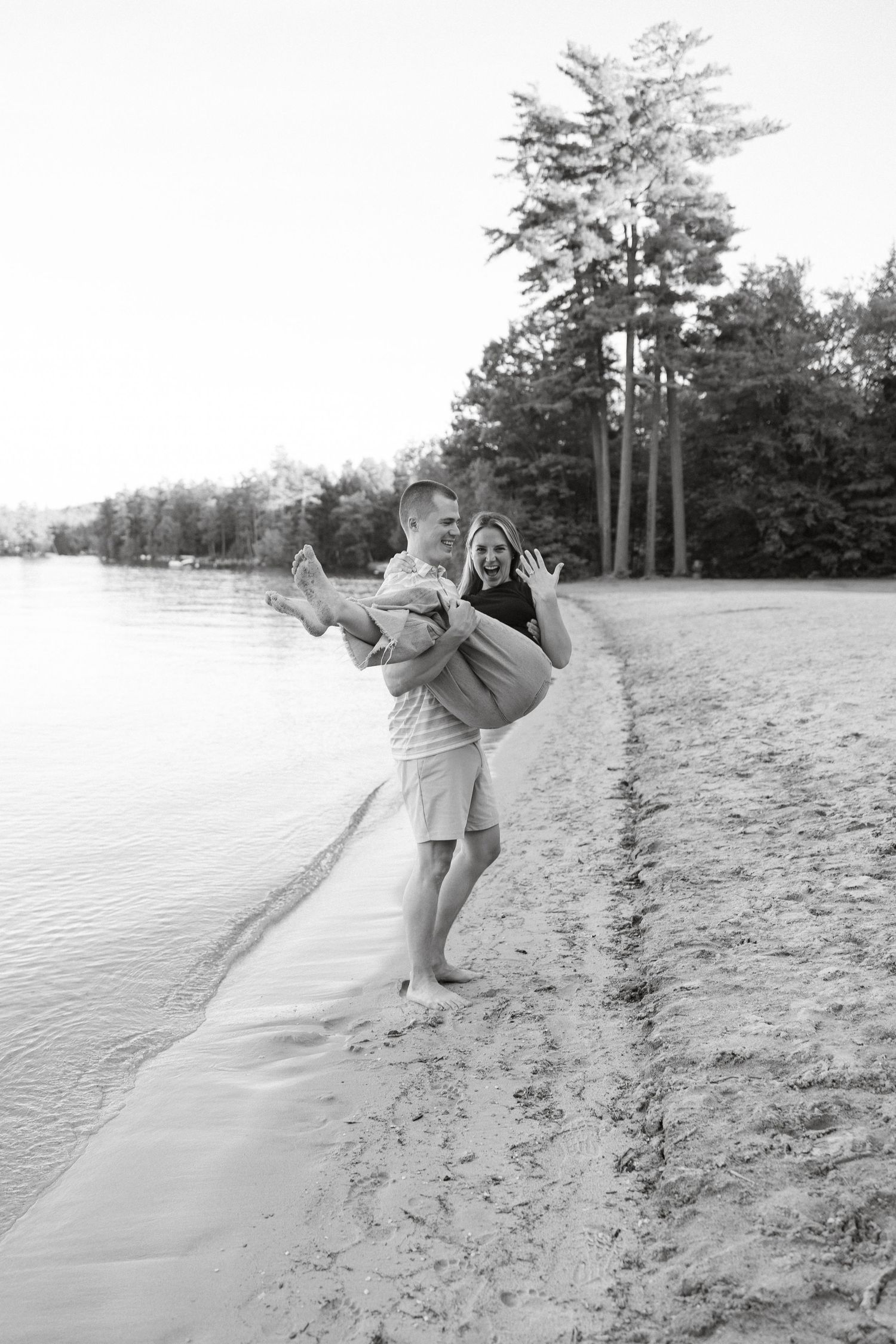 Black and white photo of a man carrying a laughing woman on a sandy beach shoreline with trees in the background.
