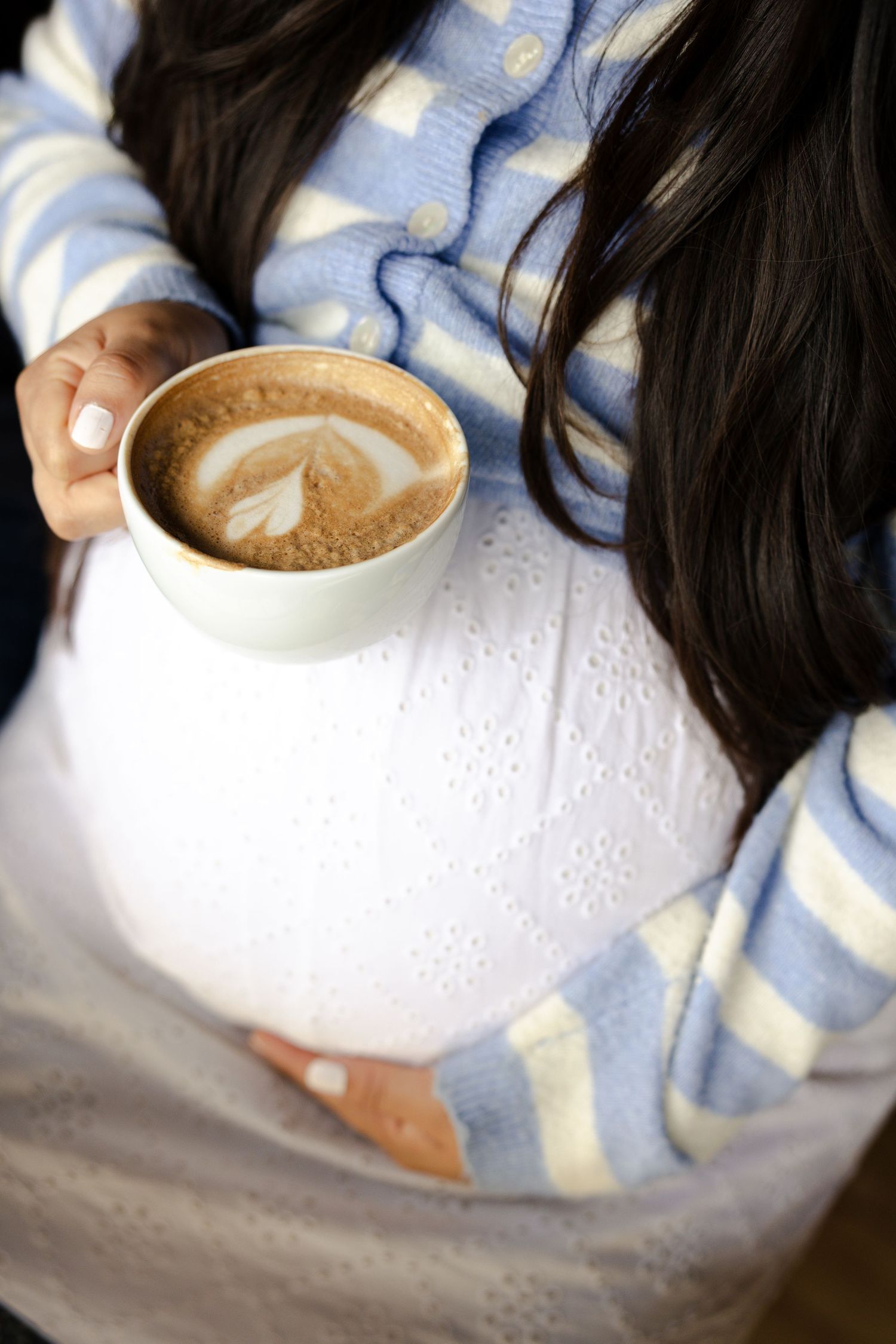 Pregnant woman in blue striped cardigan holding a latte art cappuccino cup, resting it on her baby bump.