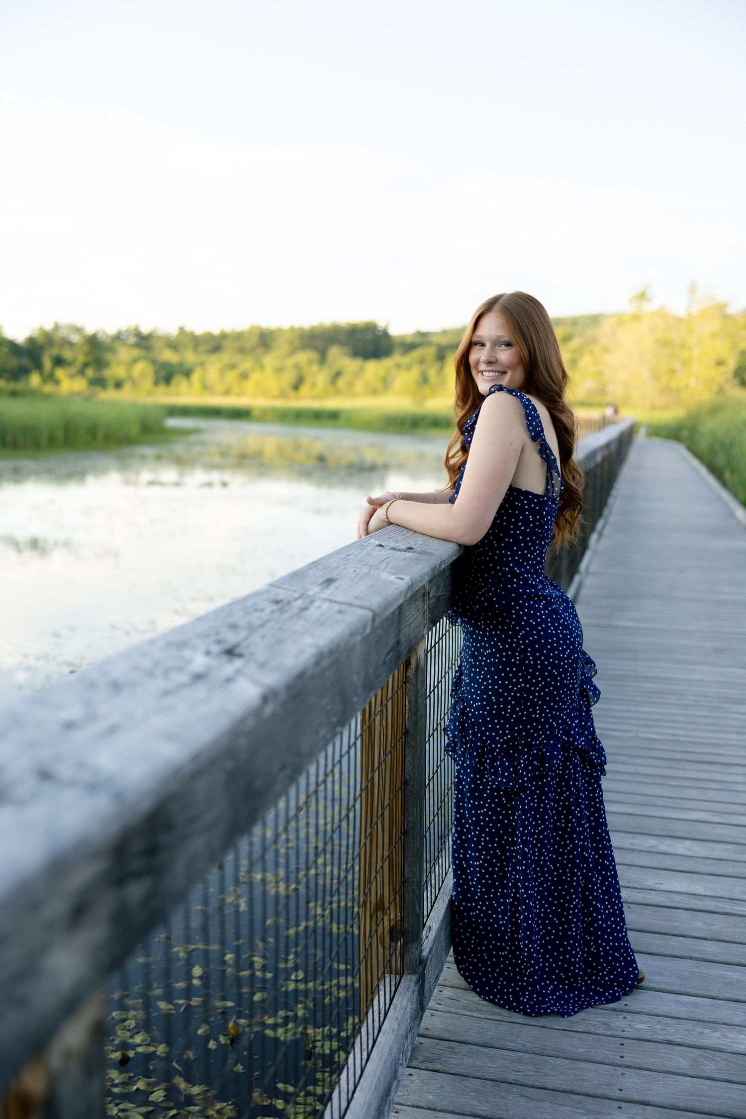Young woman in navy polka dot maxi dress smiling on wooden boardwalk bridge over scenic lake at sunset.