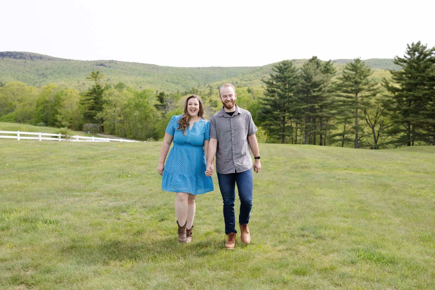 Couple smiling and holding hands while walking on green grass with trees and rolling hills in background.