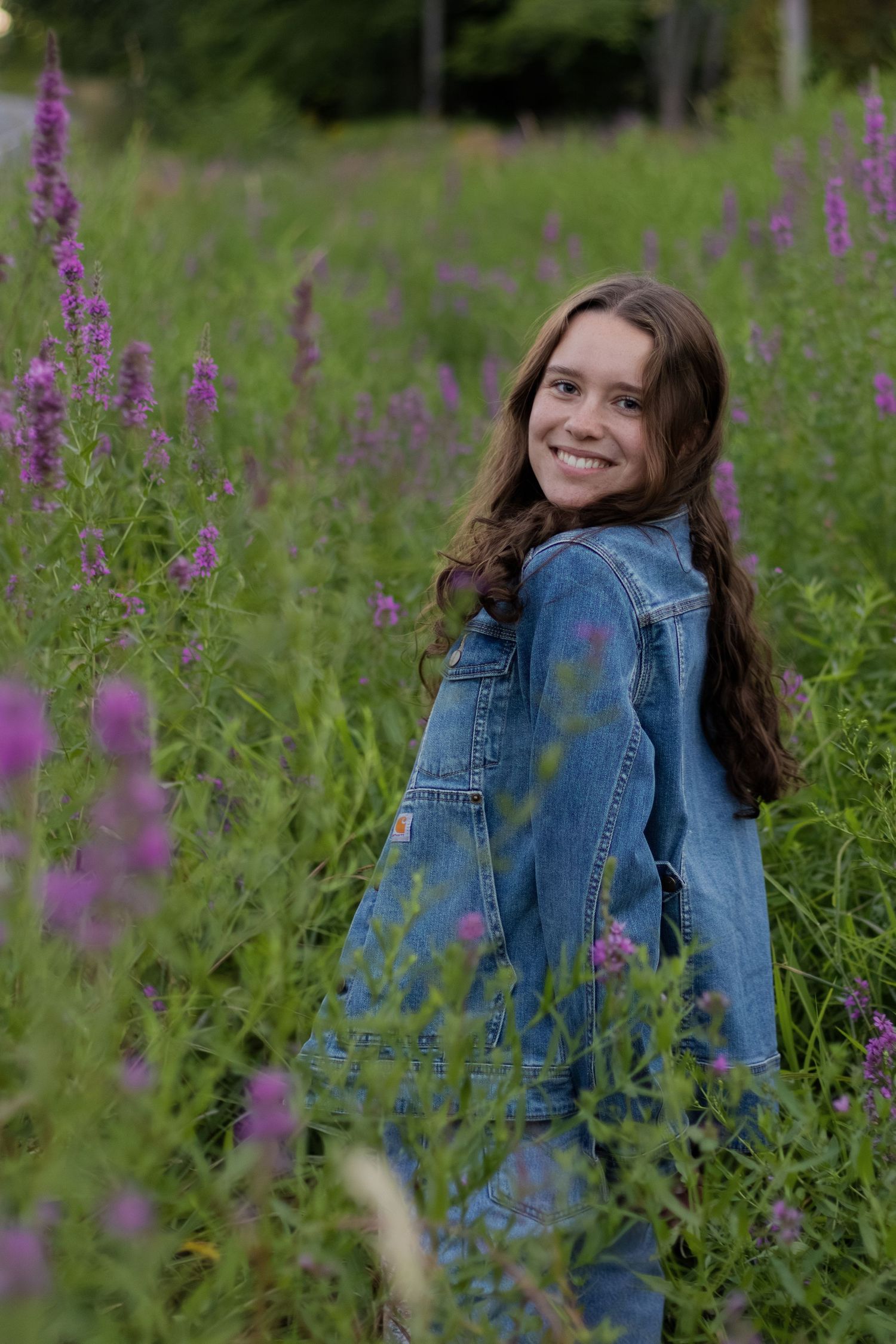 Smiling teenage girl in denim jacket sitting among purple wildflowers in a lush green field.