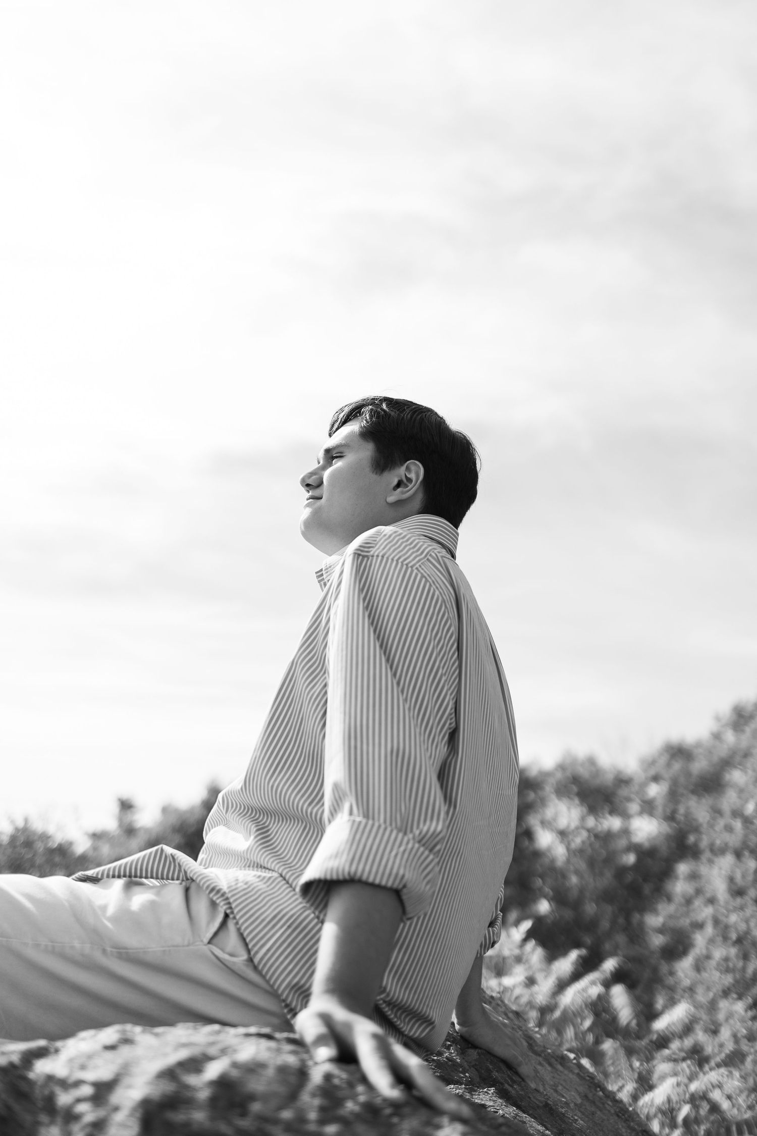 Black and white portrait of a young man in striped shirt sitting on rocks, smiling against cloudy sky.