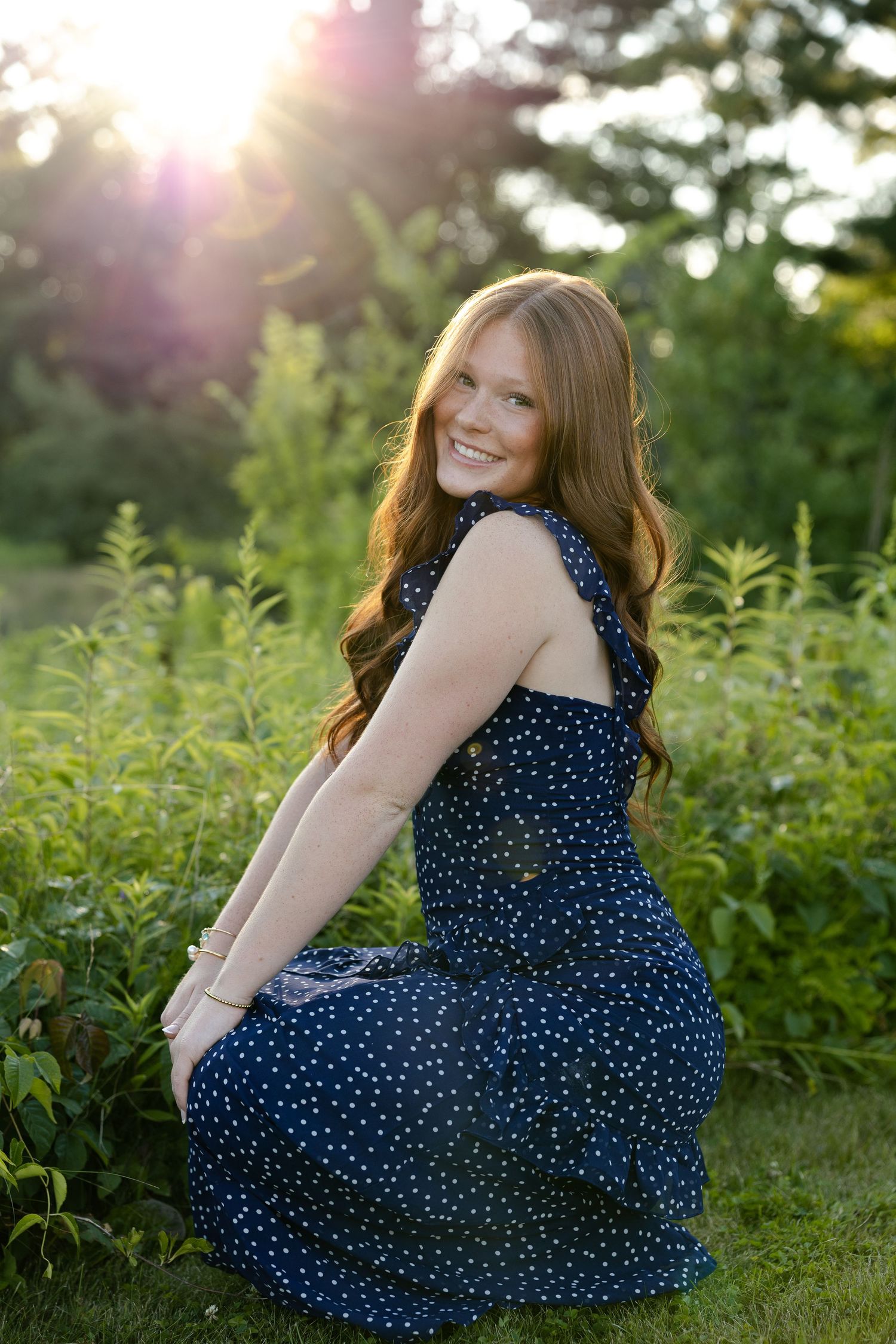 Young woman with long red hair smiling in a navy polka dot dress, seated in a lush green garden at golden hour.