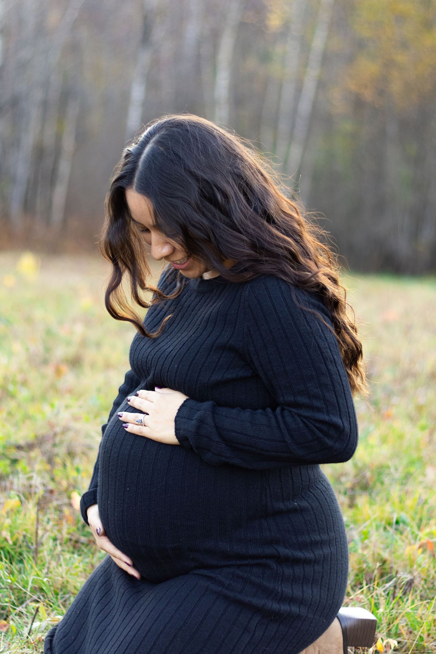 Pregnant woman in black dress cradling her baby bump outdoors in a field with autumn trees in the background.