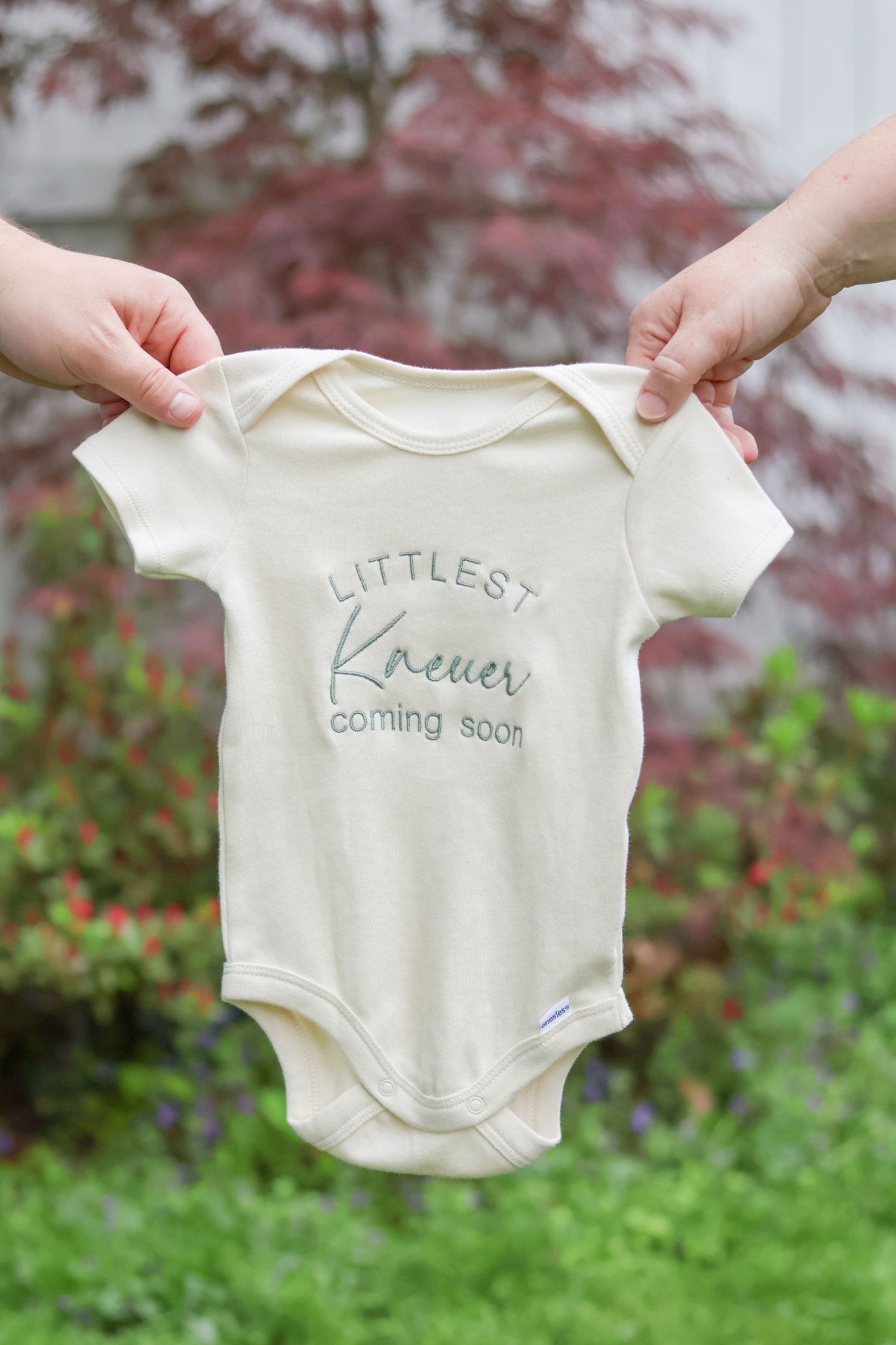 Hands holding a cream baby onesie reading 