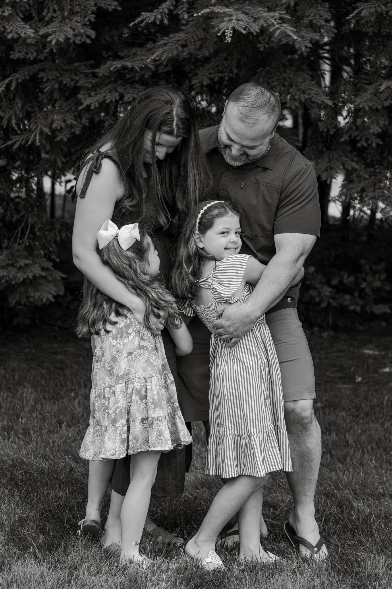 A black and white outdoor family portrait showing a loving moment together on a grassy lawn with evergreen trees.