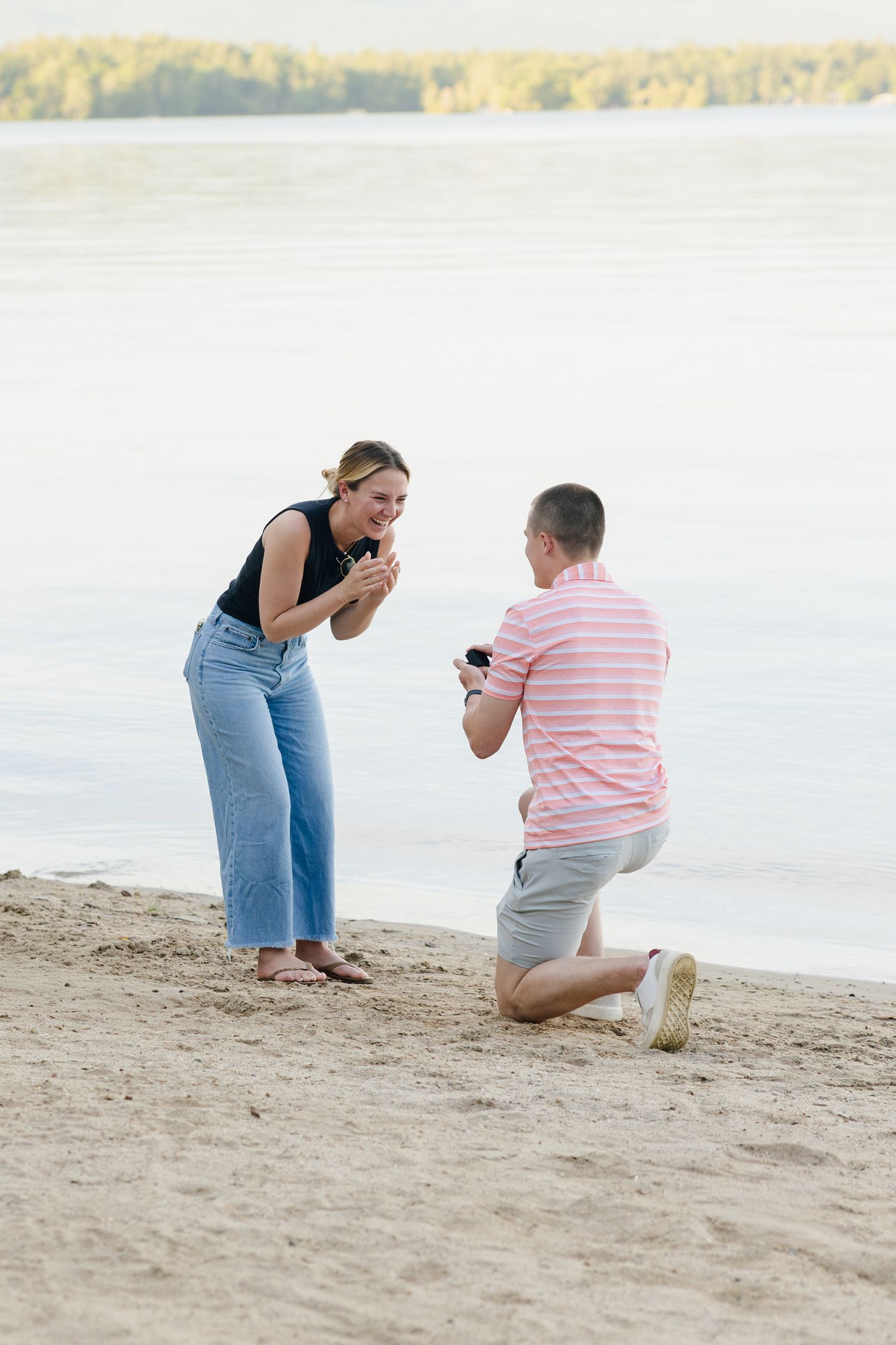 Man proposes on one knee to surprised woman on sandy beach near lake at sunset.