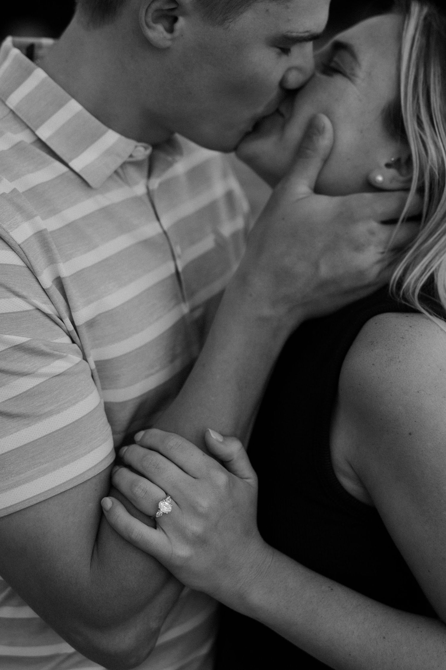 Black and white photo of a couple kissing, woman displaying diamond engagement ring, romantic proposal moment.
