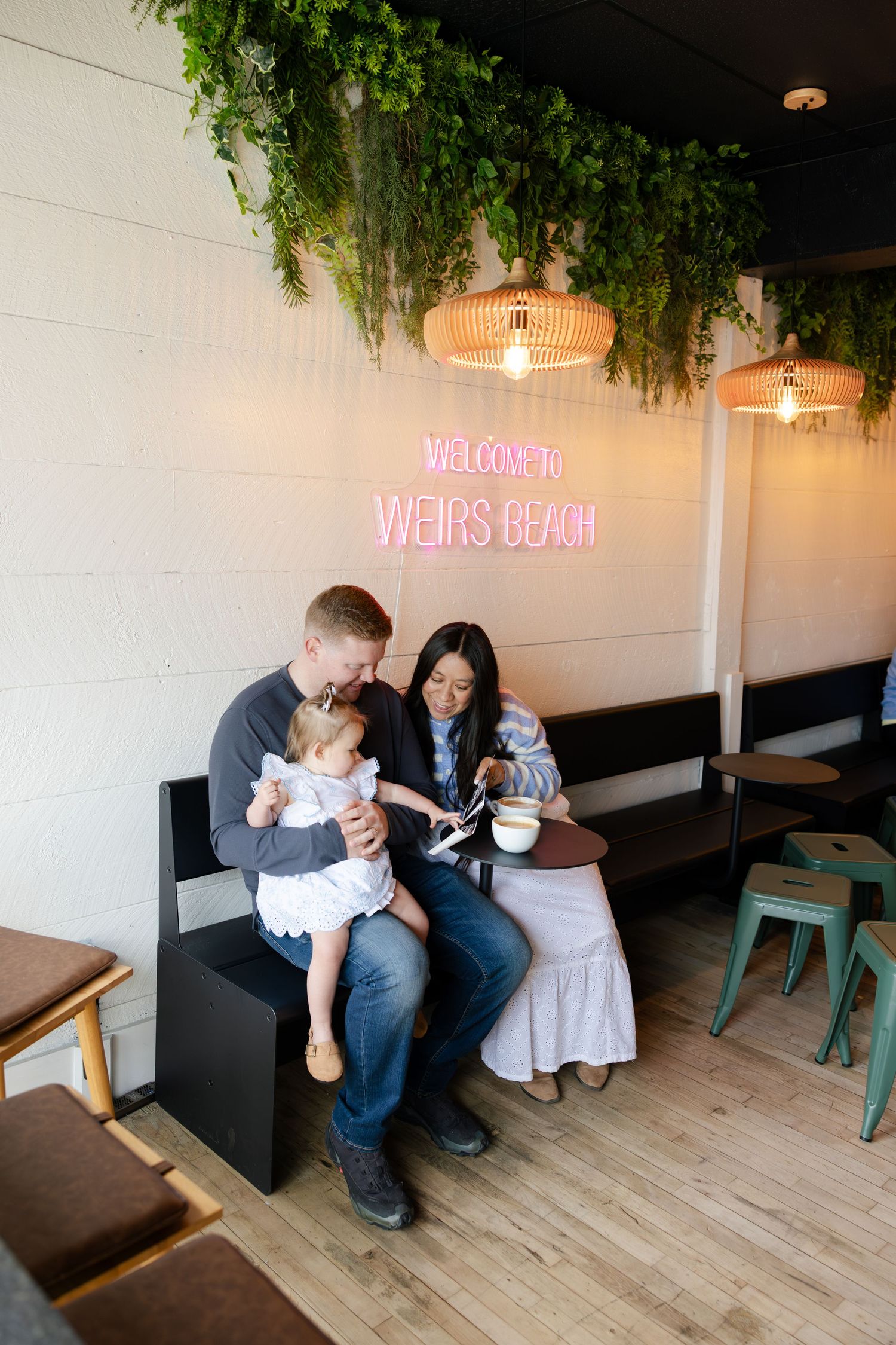 Family enjoying ice cream together on a bench inside Weirs Beach cafe with neon sign and greenery decor.