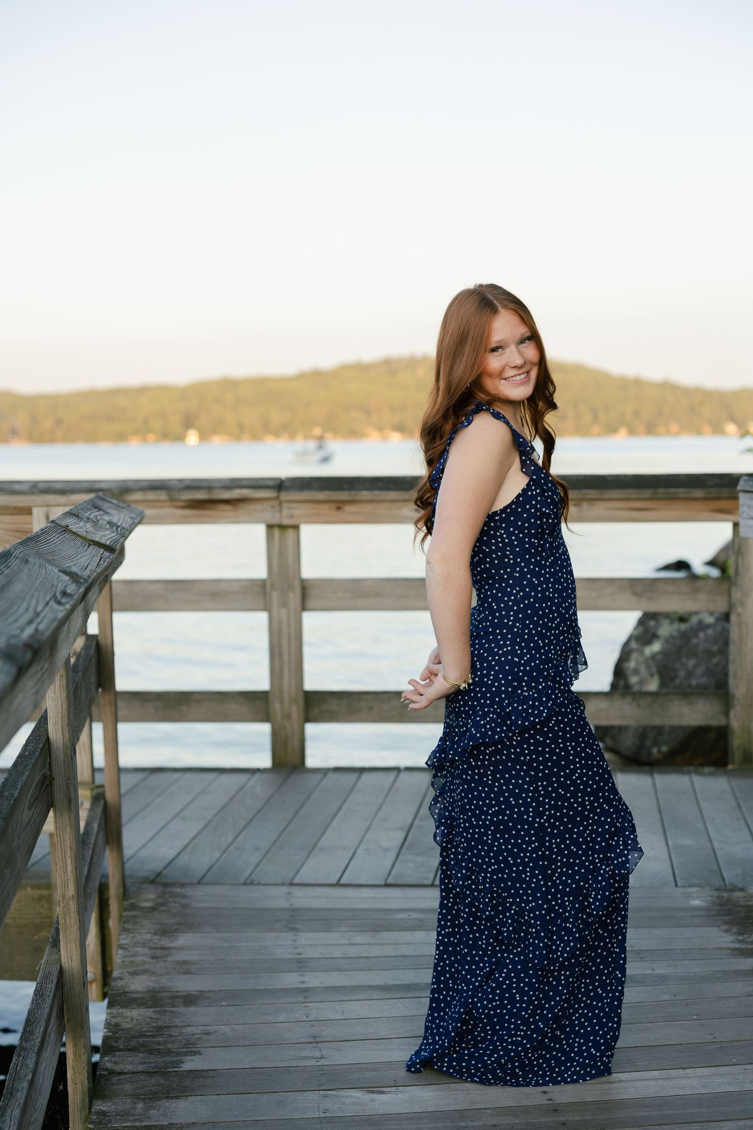 Young woman in navy blue polka dot maxi dress smiling on wooden pier with water and hills in background.