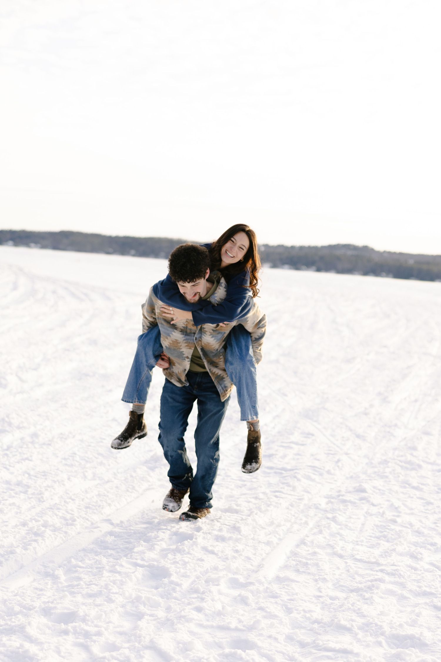 A man gives a woman a piggyback ride across a snowy field in winter, both smiling and dressed in casual cold-weather clothing.