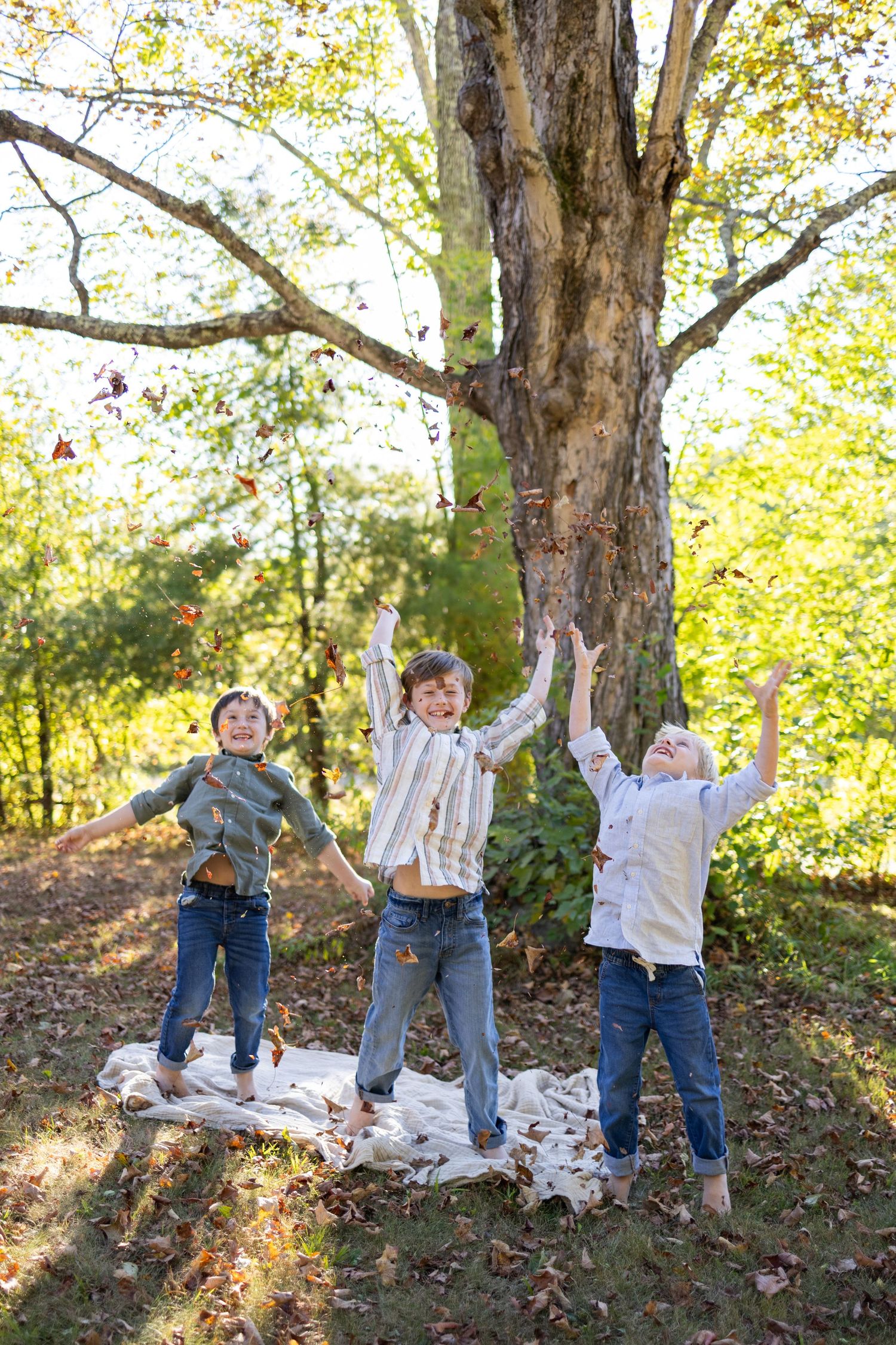 Children playing and jumping among fall leaves in a wooded area wearing casual attire.