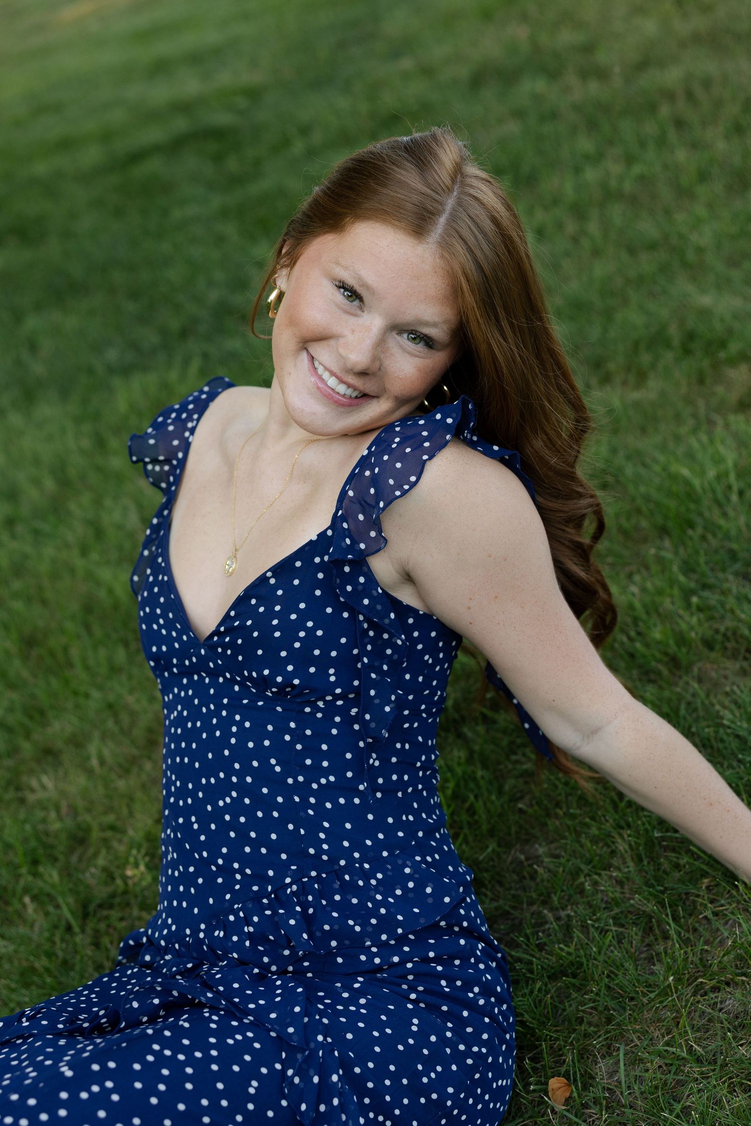 Smiling redhead woman in navy polka dot dress sitting on green grass, looking up cheerfully.