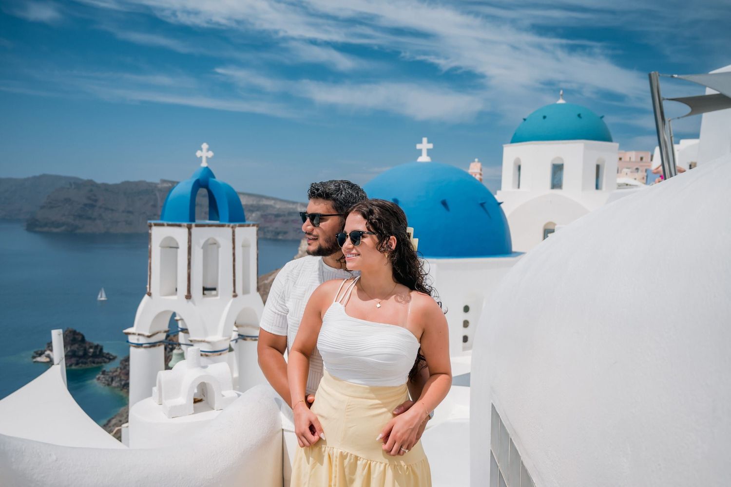 A couple poses on whitewashed steps with Santorini's colorful architecture and blue domes in the background.