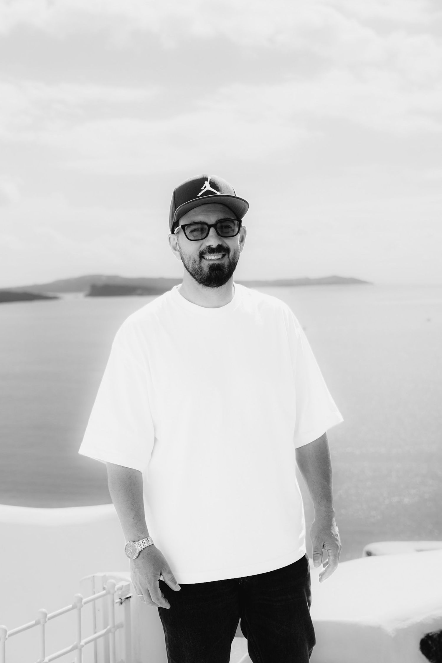 Man in white oversized tee and Jordan snapback smiling with sunglasses, posing against scenic ocean backdrop.