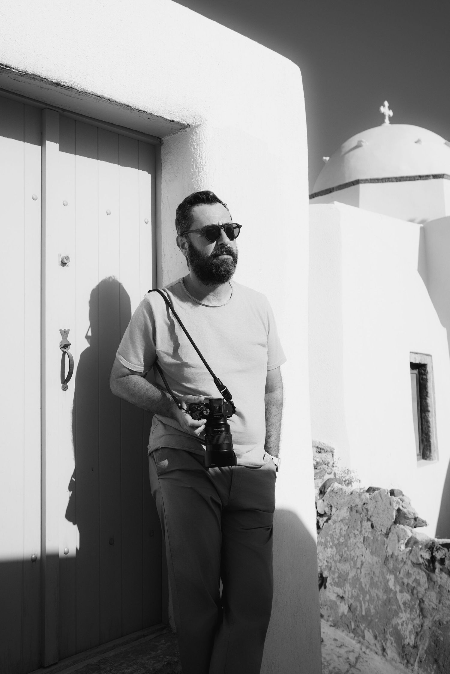 Bearded man with sunglasses holding a camera leaning against a white wall near a church in Santorini, black and white photo.