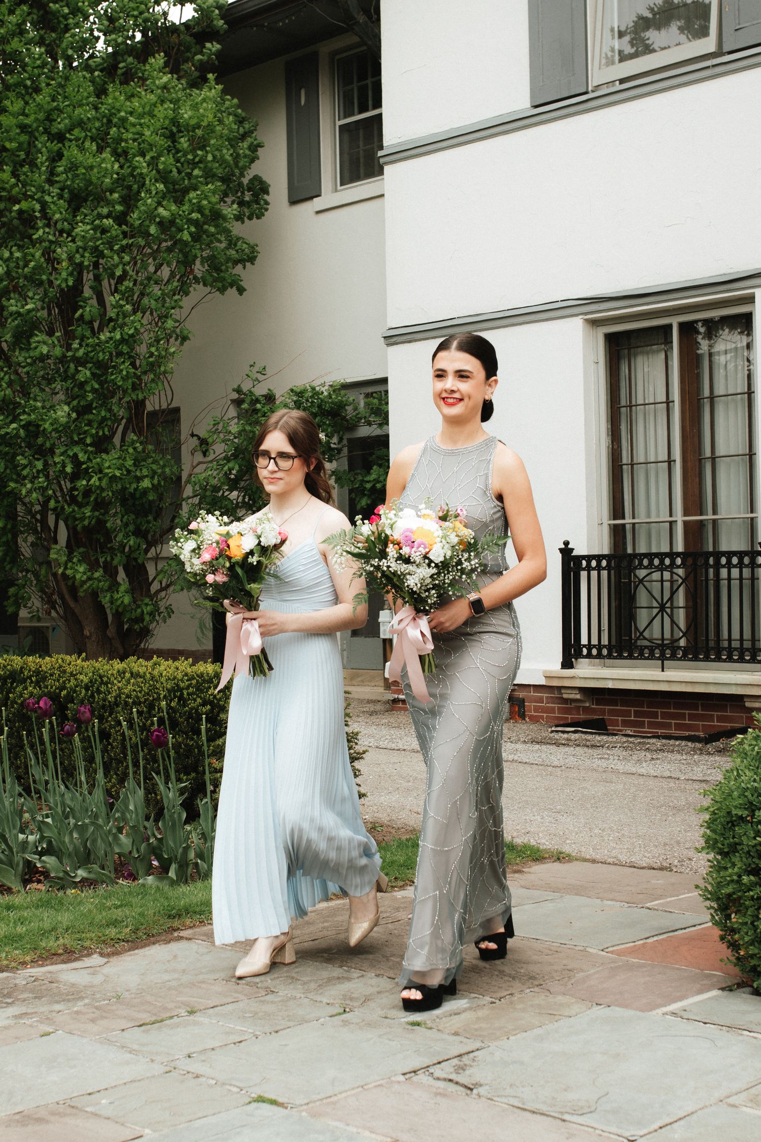 Wedding party members in light blue and gray dresses hold bouquets while walking outside an elegant white building.