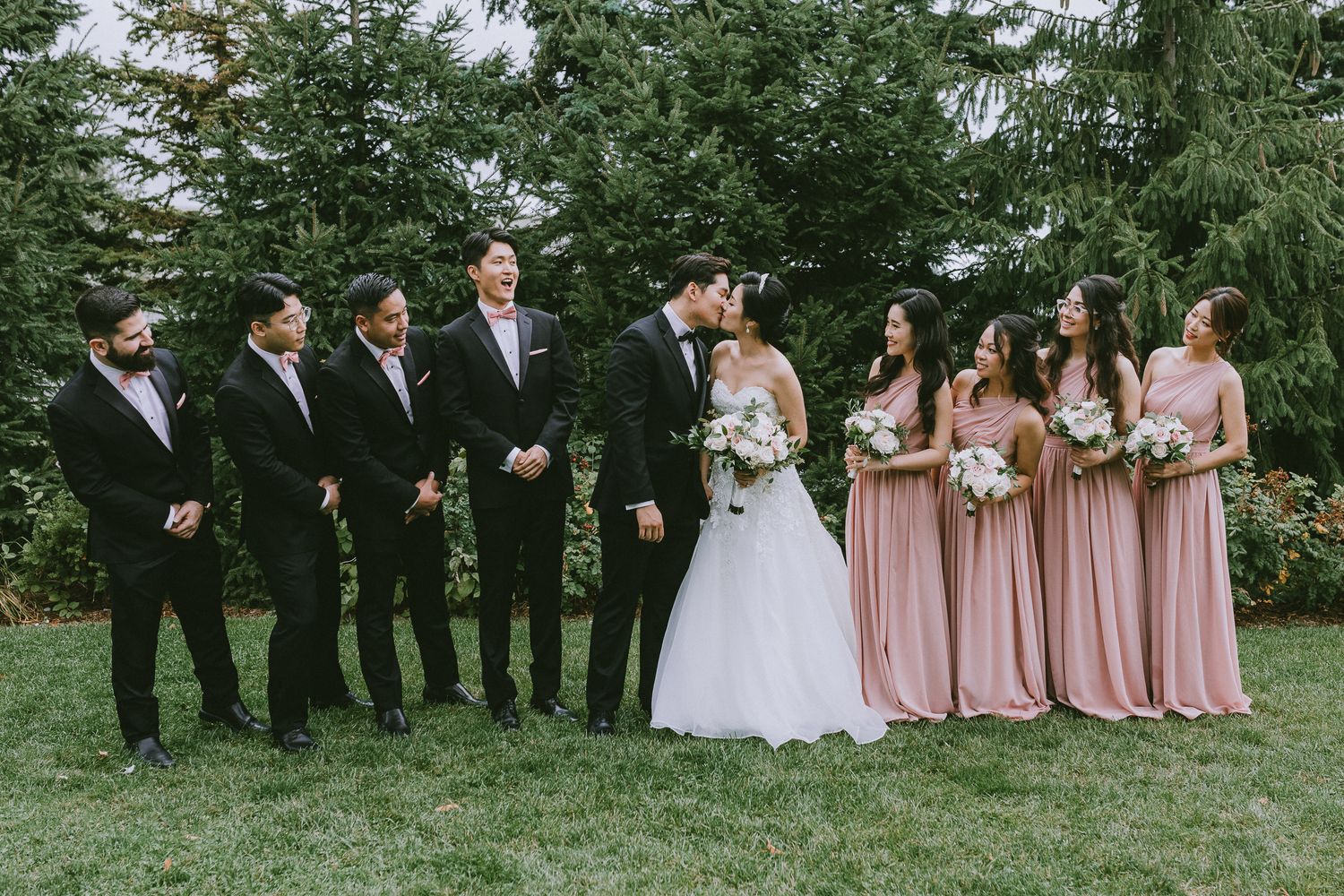 Wedding party in formal attire poses outdoors with bridesmaids in pink dresses and groomsmen in dark suits.