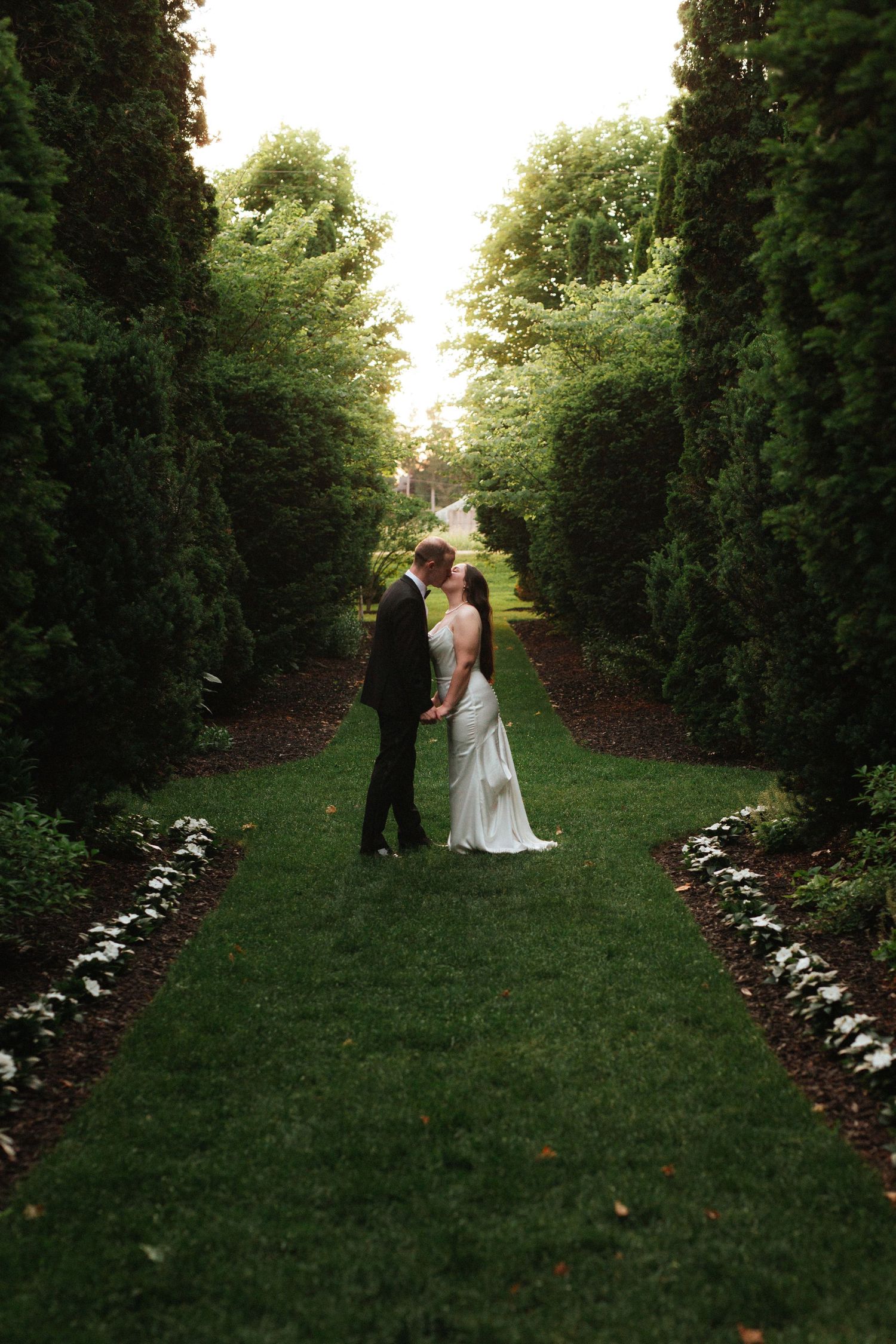 Bride and groom sharing a romantic moment on a lush green garden path lined with tall hedges and trees.