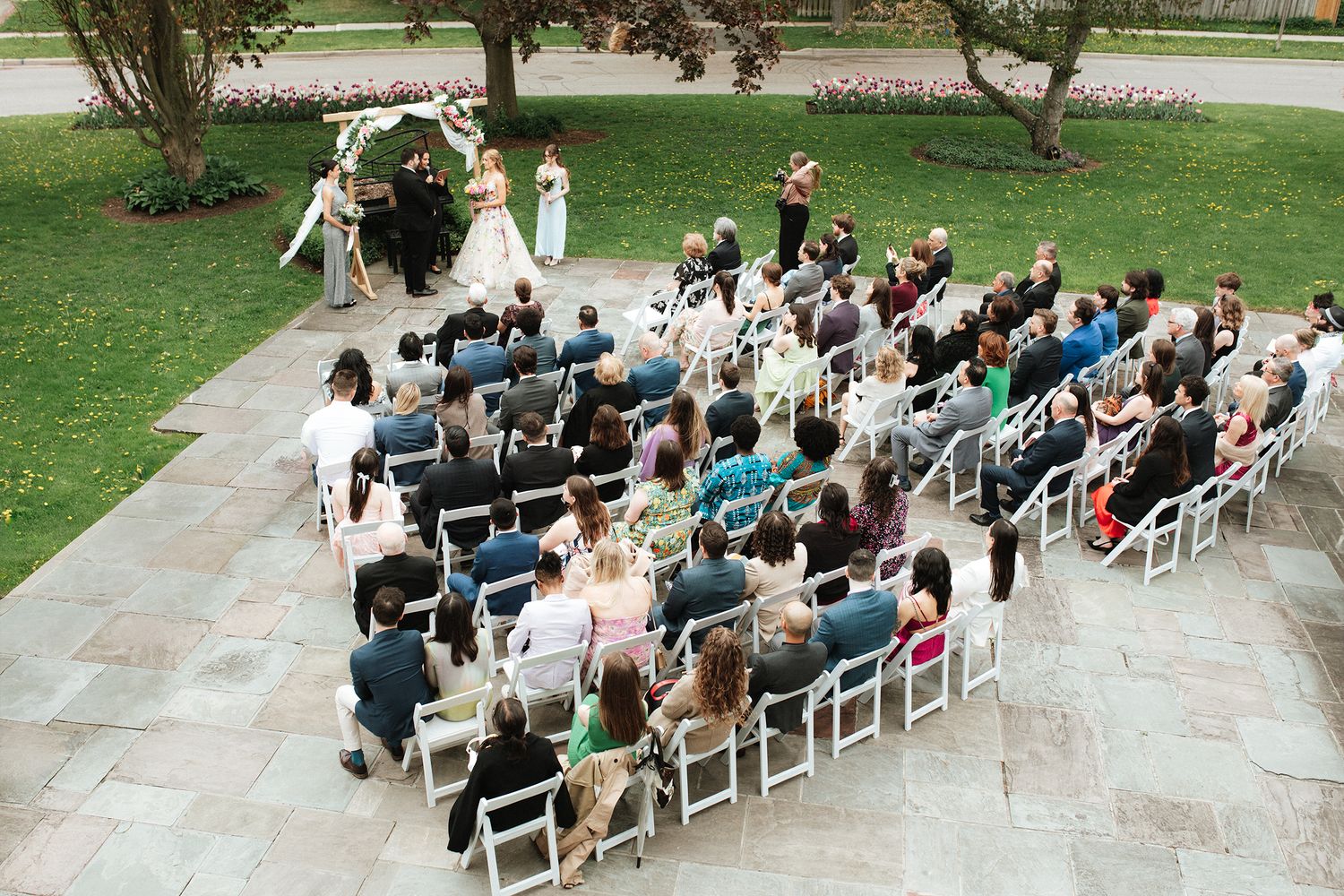 Outdoor wedding ceremony with guests seated in white chairs on stone patio surrounded by green lawn and trees.