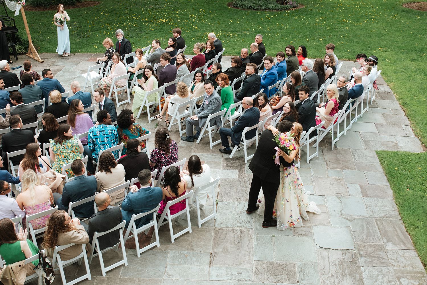 Aerial view of an outdoor wedding ceremony with guests seated in white chairs on a stone patio surrounded by green lawn.