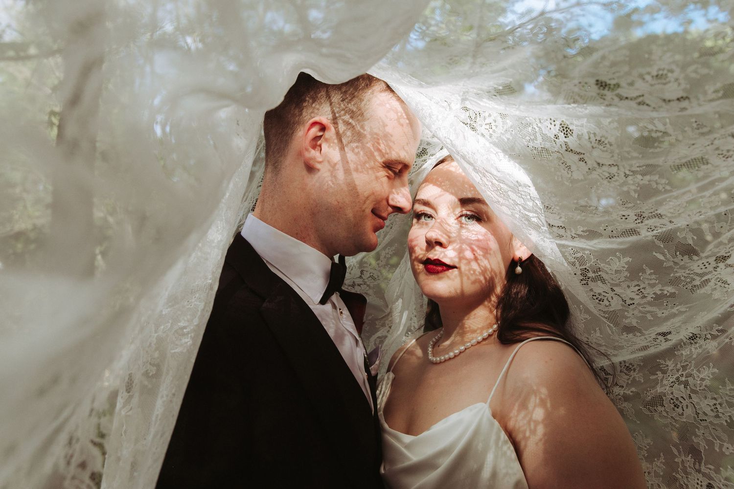 Bride and groom share an intimate moment under a flowing lace wedding veil, gazing into each other's eyes.