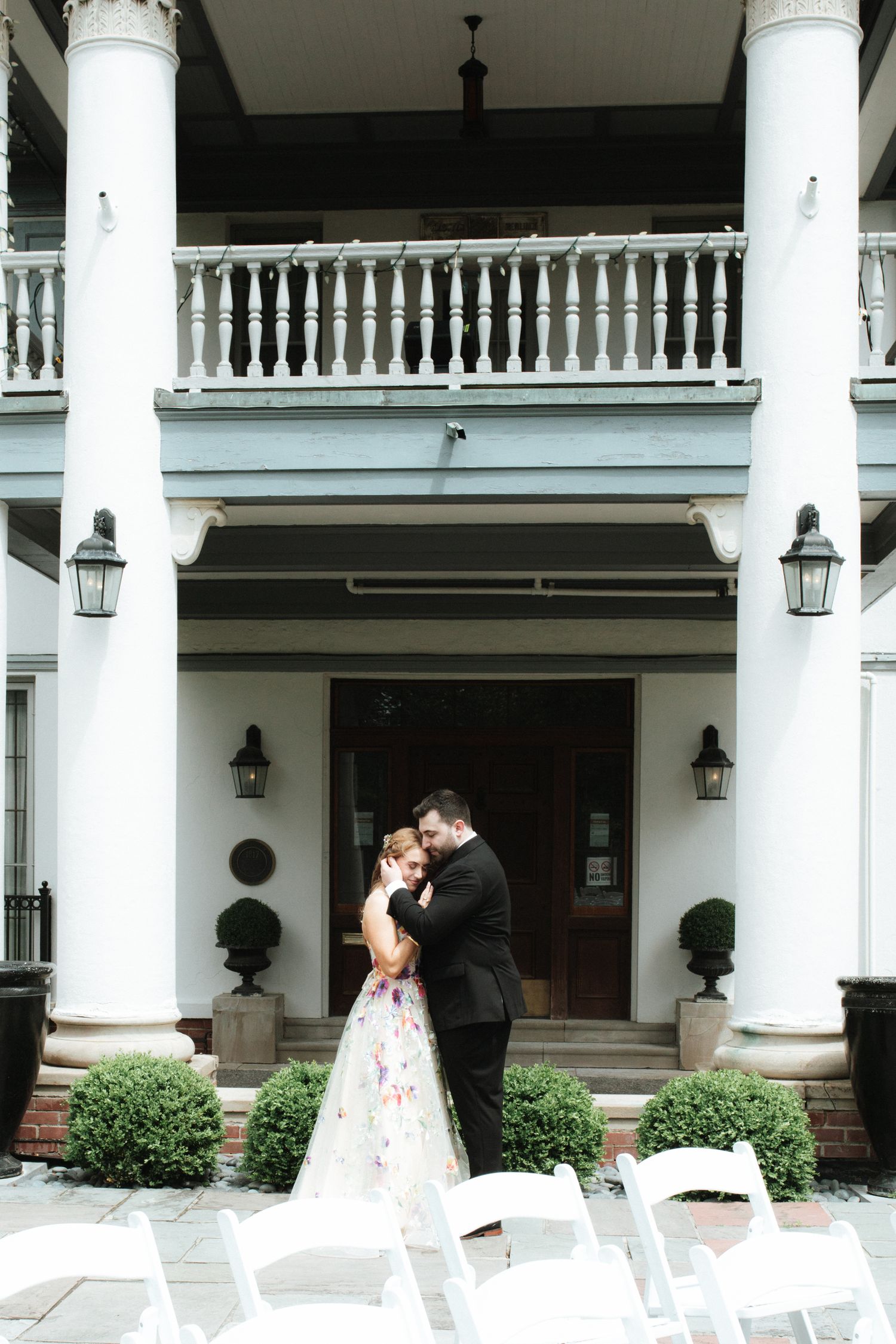 A couple shares a romantic moment on their wedding day in front of a grand white columned entrance with chairs set up.