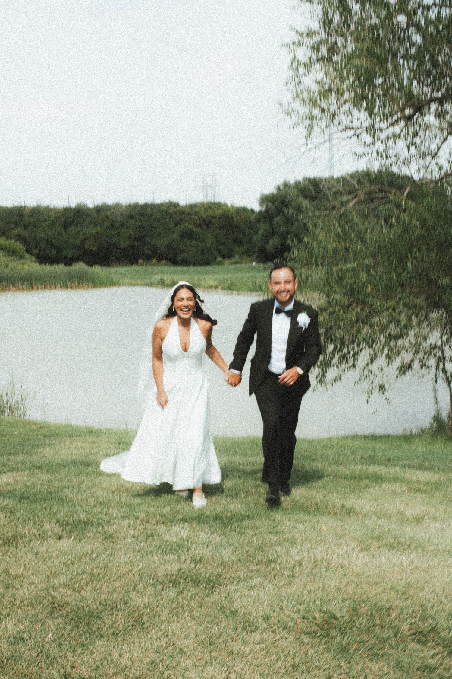 A couple in wedding attire runs playfully across a grassy field near a lake on their special day.