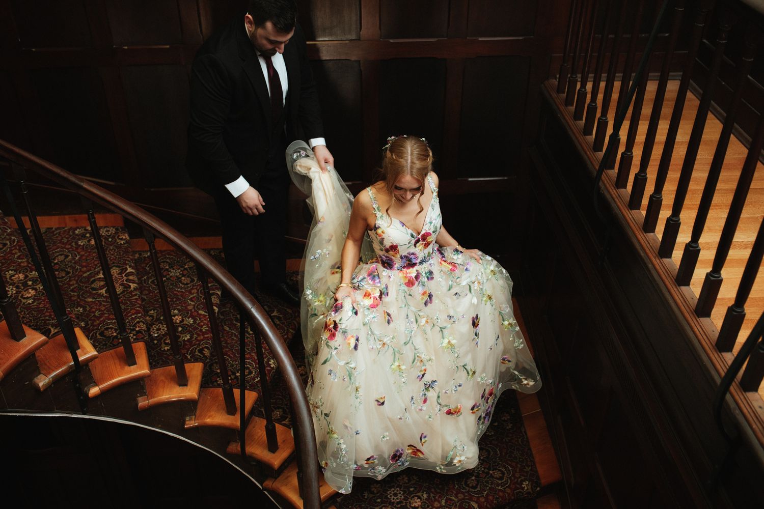A person in a floral dress descends a wooden staircase in a dimly lit interior space.