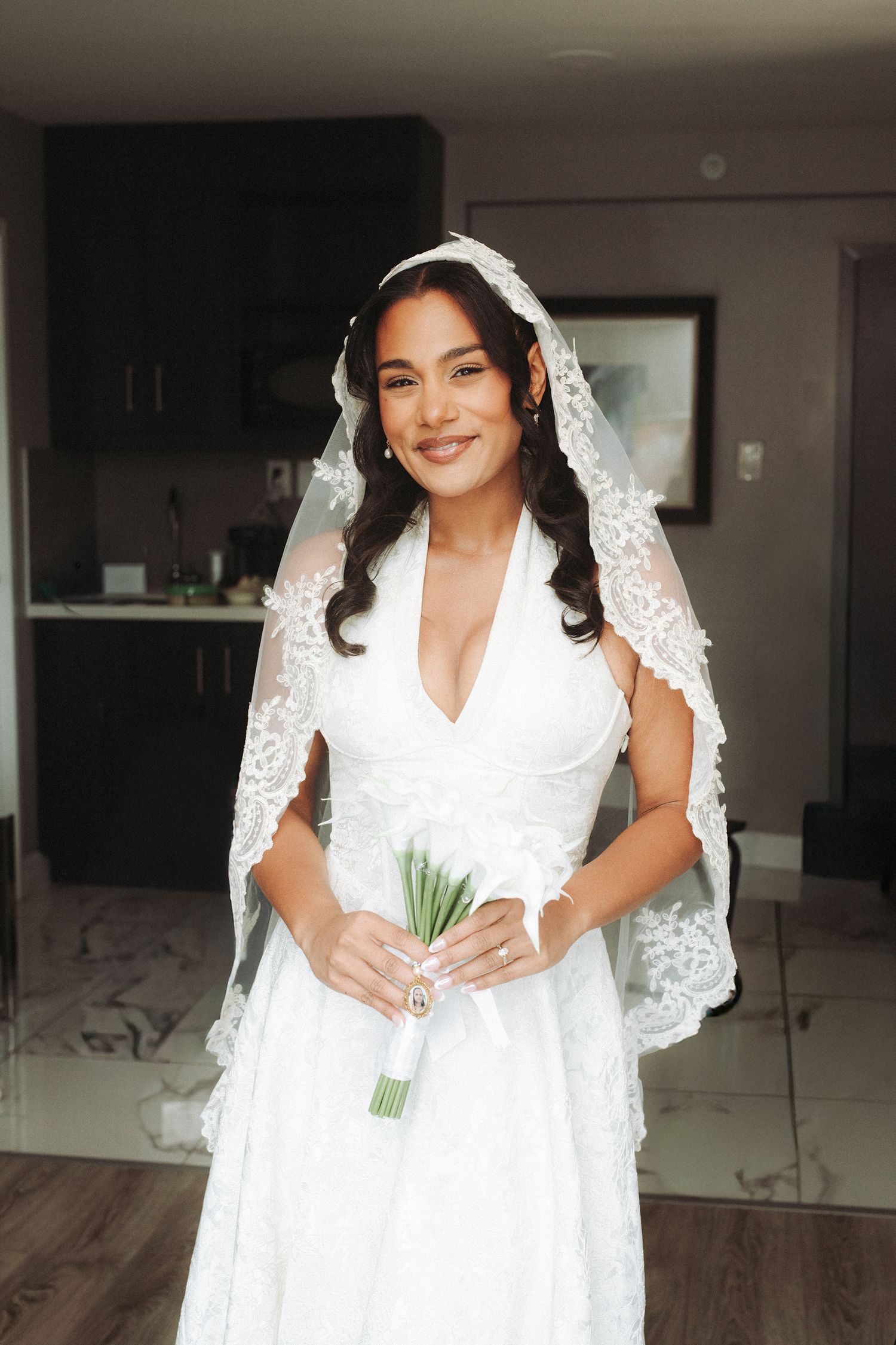 A bride in a white wedding dress and lace veil holds a small bouquet while getting ready in an indoor setting.