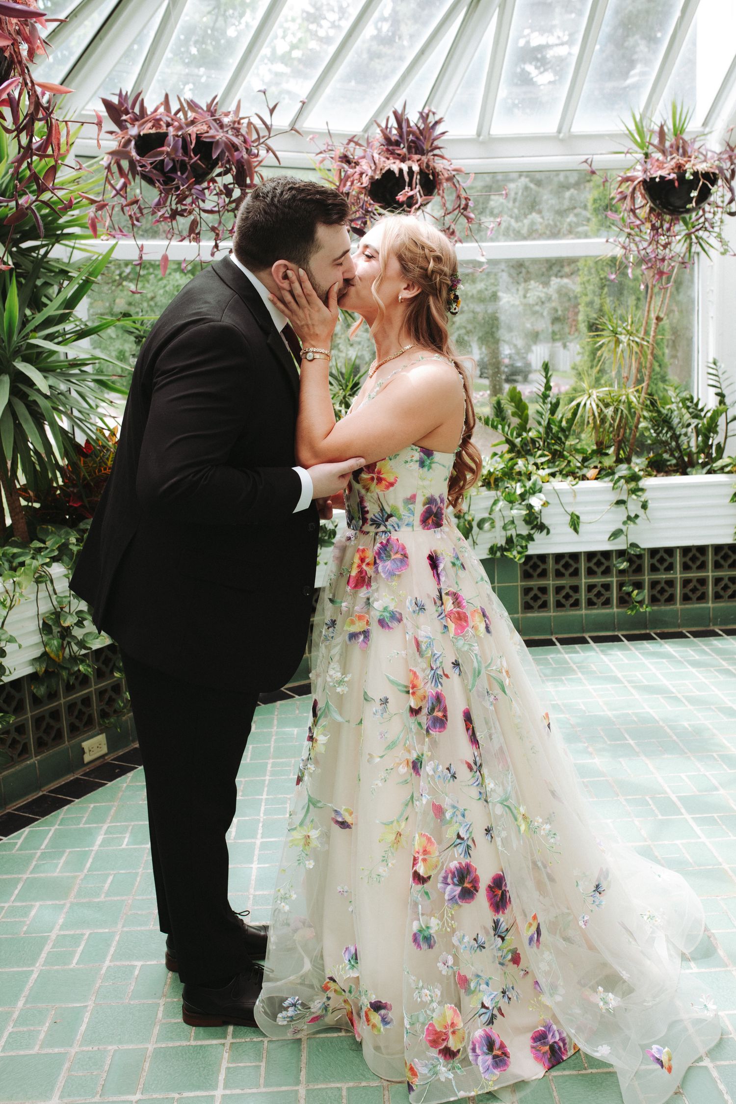 A romantic wedding moment in a greenhouse with a floral dress and black tuxedo surrounded by tropical plants and flowers.