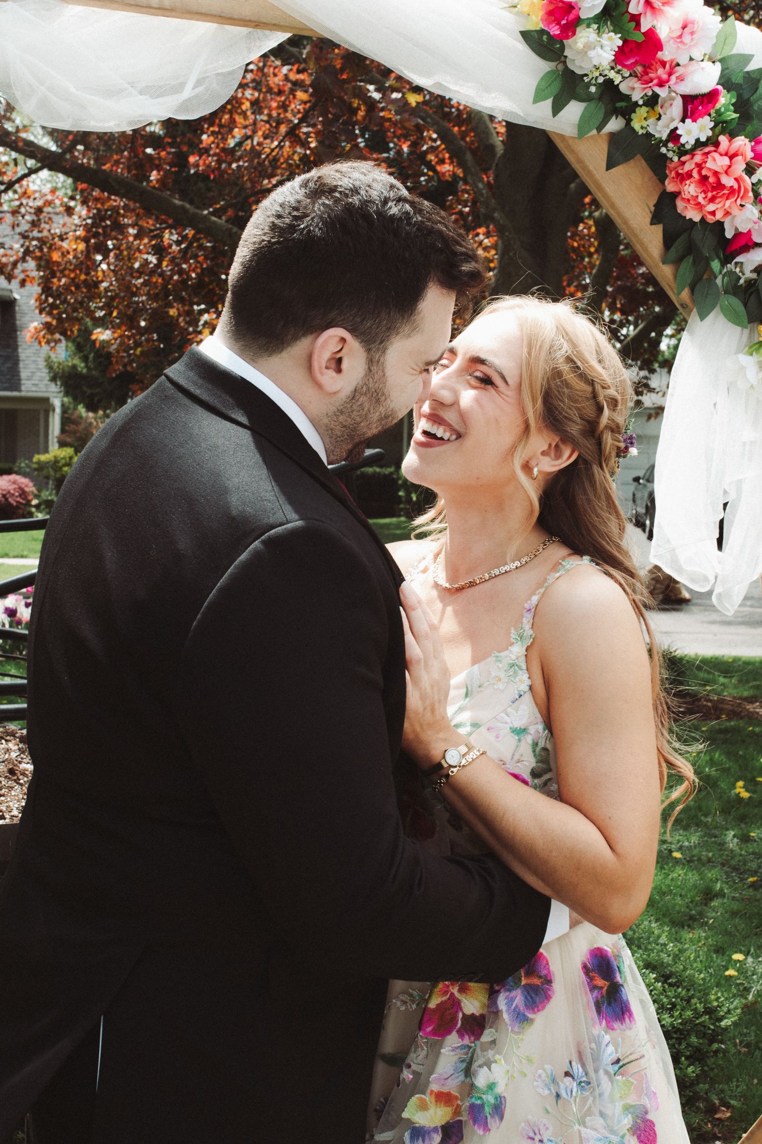 A couple shares a joyful moment in a garden setting, with the person wearing a floral dress laughing during their embrace.