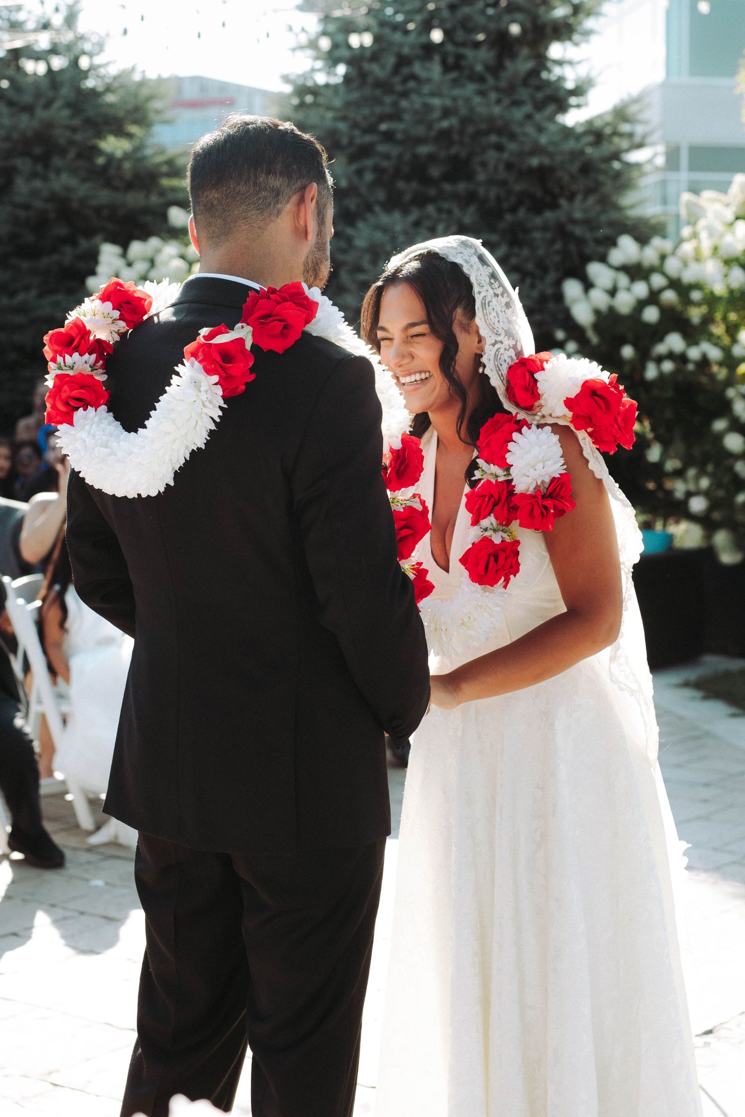 A joyful wedding couple wearing traditional Hawaiian red and white leis share a candid moment outdoors.