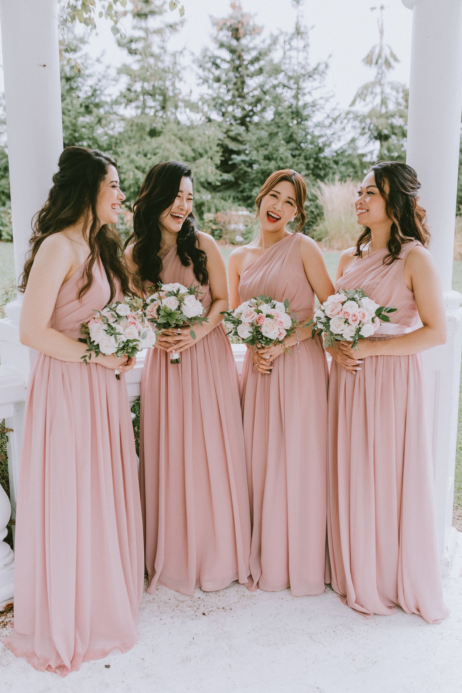 Bridesmaids in blush pink floor-length dresses share a candid moment while holding pink and white bouquets.