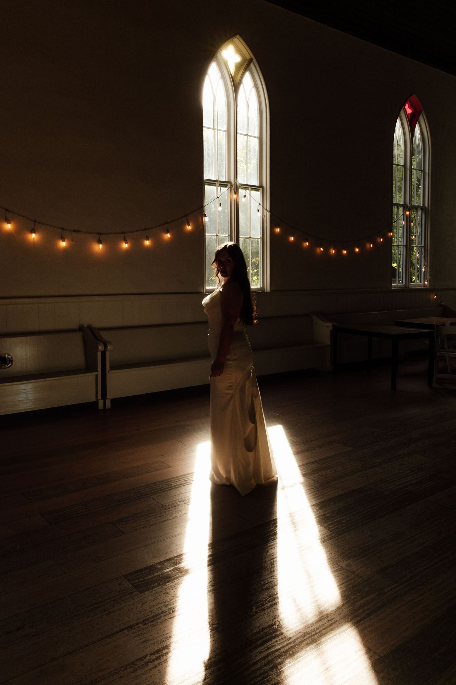 Bride in elegant white gown bathed in dramatic window light inside a warmly lit vintage church hall.