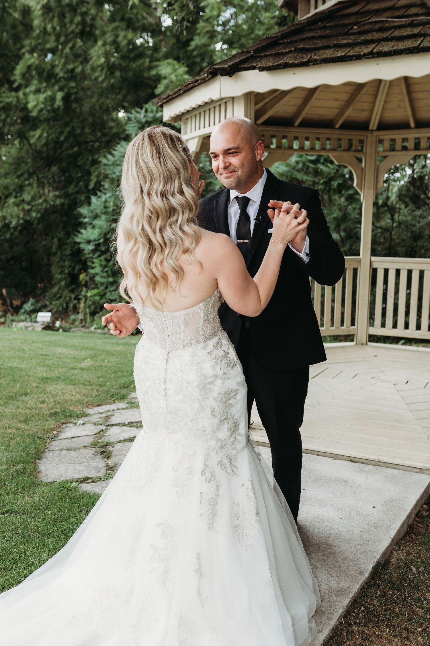 A couple in formal wedding attire dance together under a wooden gazebo surrounded by lush greenery.
