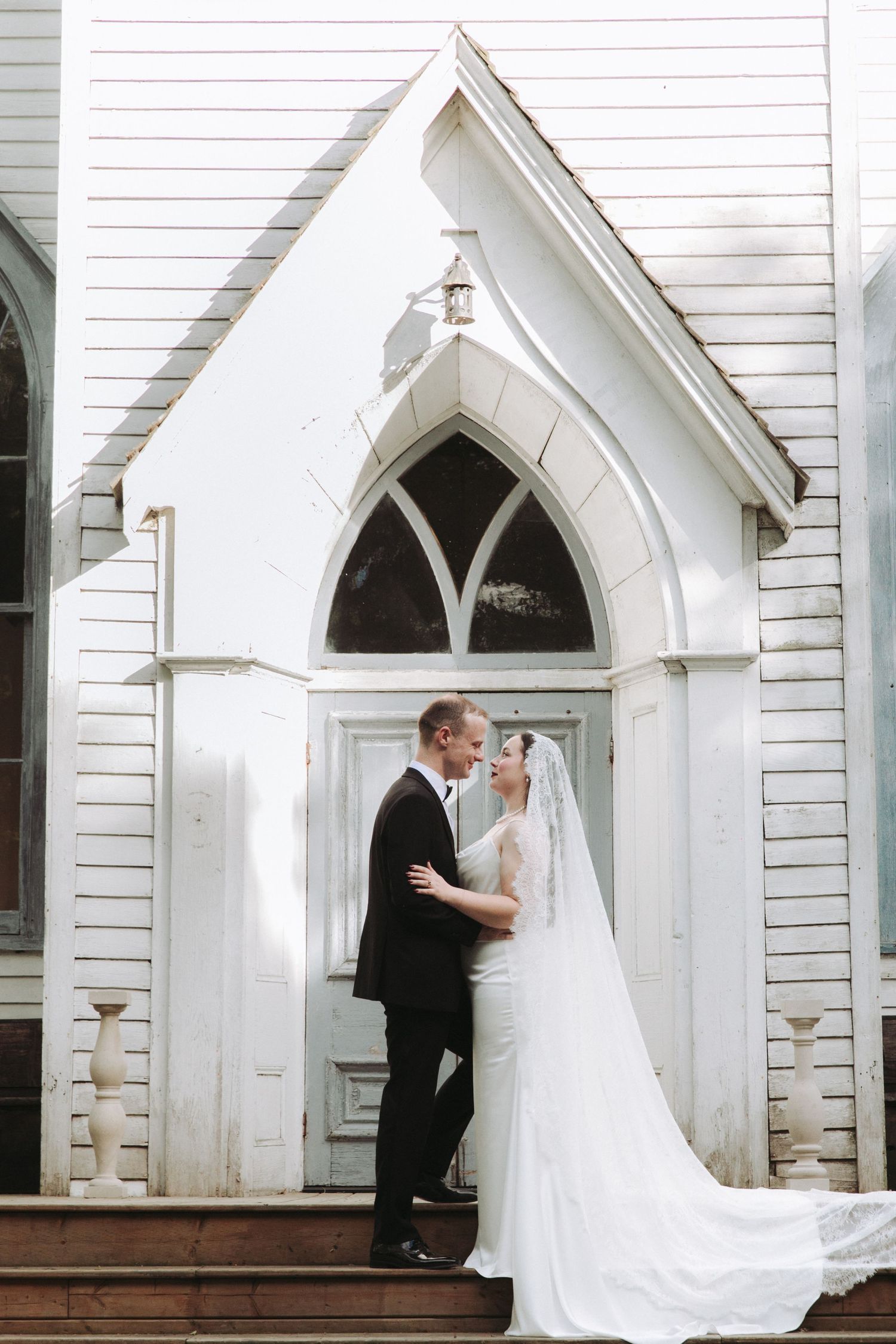 Bride and groom share a romantic moment on the steps of a white Gothic-style church on their wedding day.