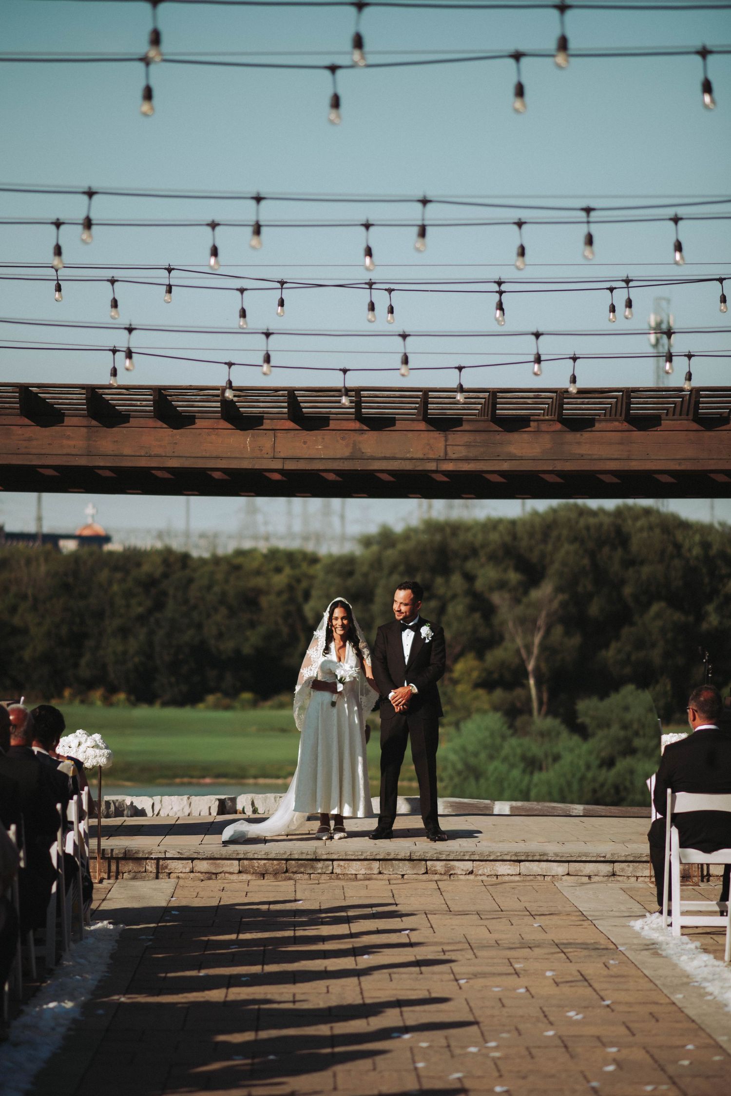 String lights hang overhead at an outdoor wedding ceremony venue with guests seated on stone pavers at sunset.