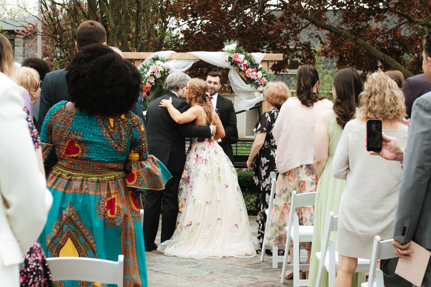 Guests celebrate at an outdoor wedding ceremony with colorful decorations and natural greenery in the background.