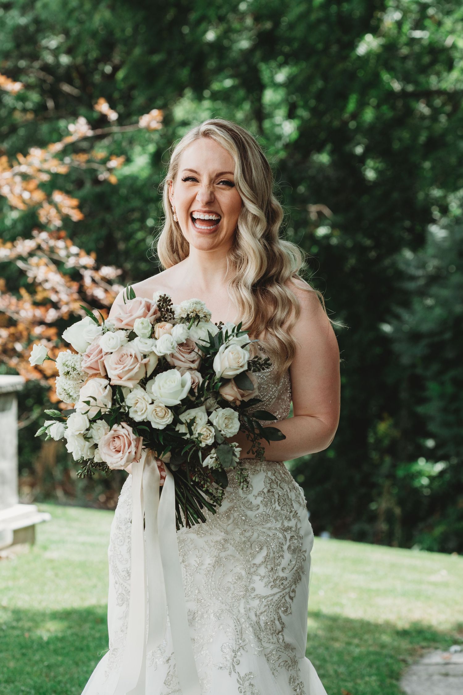 A joyful bride in a white lace wedding dress holds a romantic bouquet of blush and ivory flowers in an outdoor garden setting.