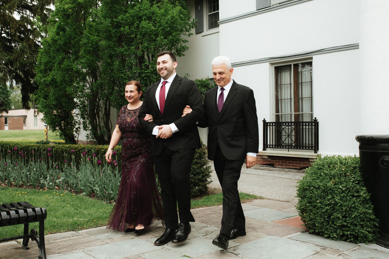 Three people walk together along a garden path outside a white building with manicured hedges.