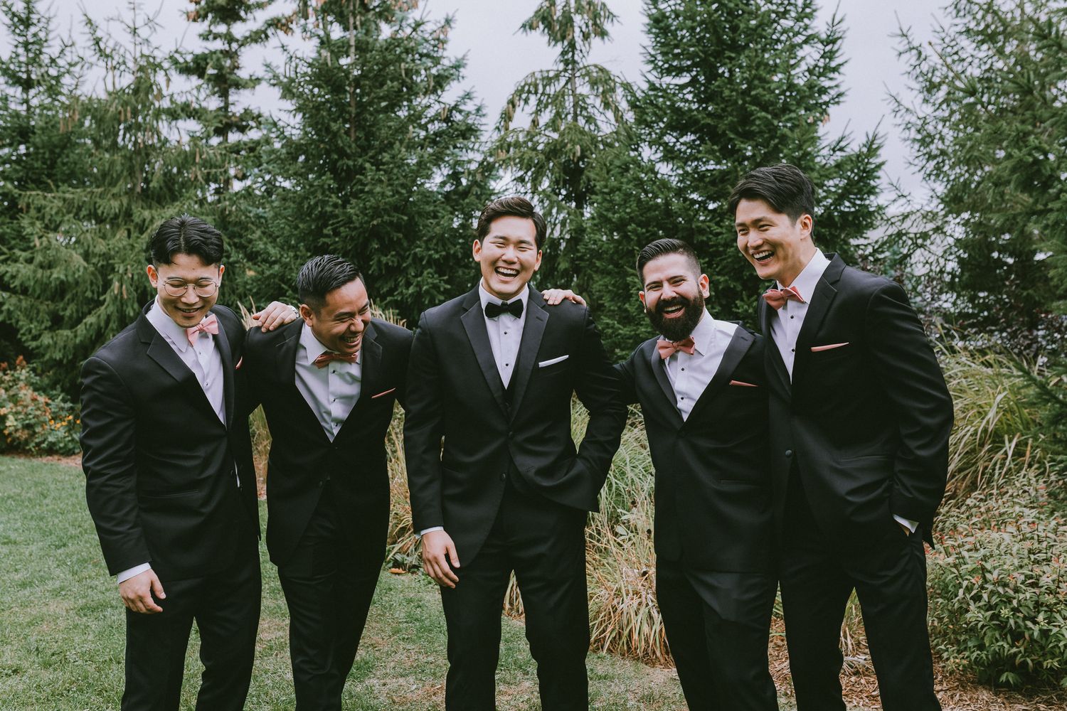 Groomsmen in black suits share a candid moment of laughter while walking outdoors on a wedding day.