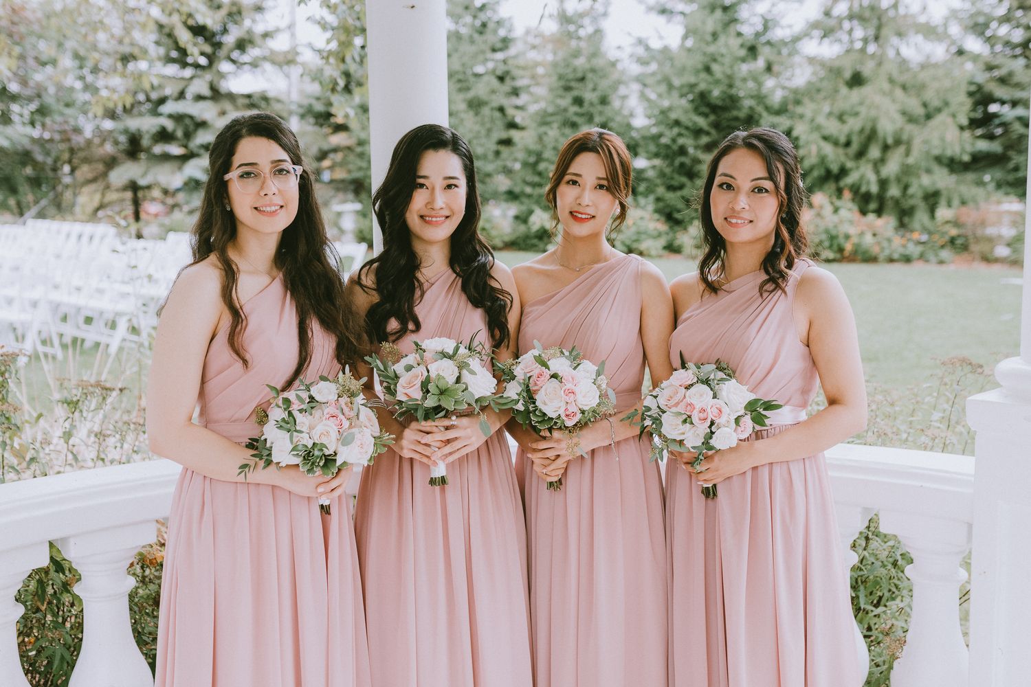 Bridesmaids in flowing pink dresses stand together holding white and pink floral bouquets at an outdoor wedding venue.